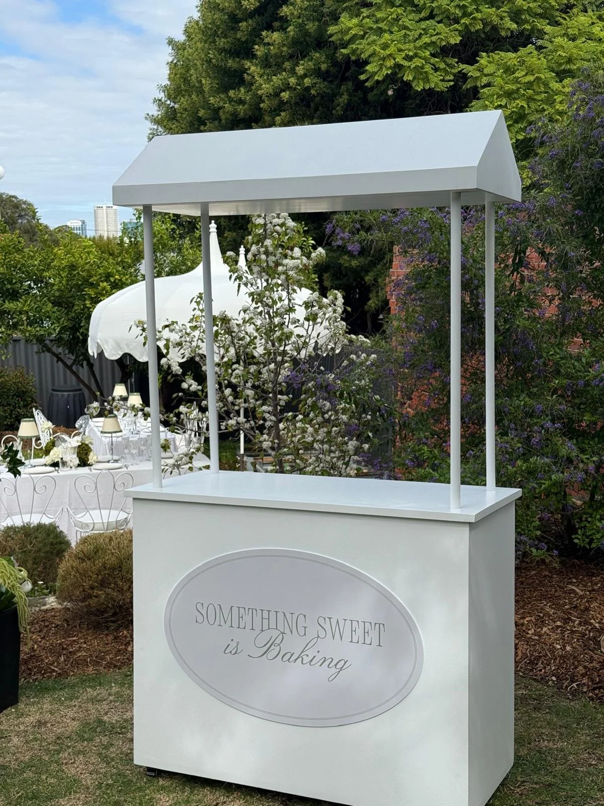 A white bakery cart with a canopy, featuring a sign that reads "Something sweet is Baking." The cart is in a garden with trees and purple and white flowering plants, and there is a white outdoor dining setup with tables, chairs, and umbrellas in the background.