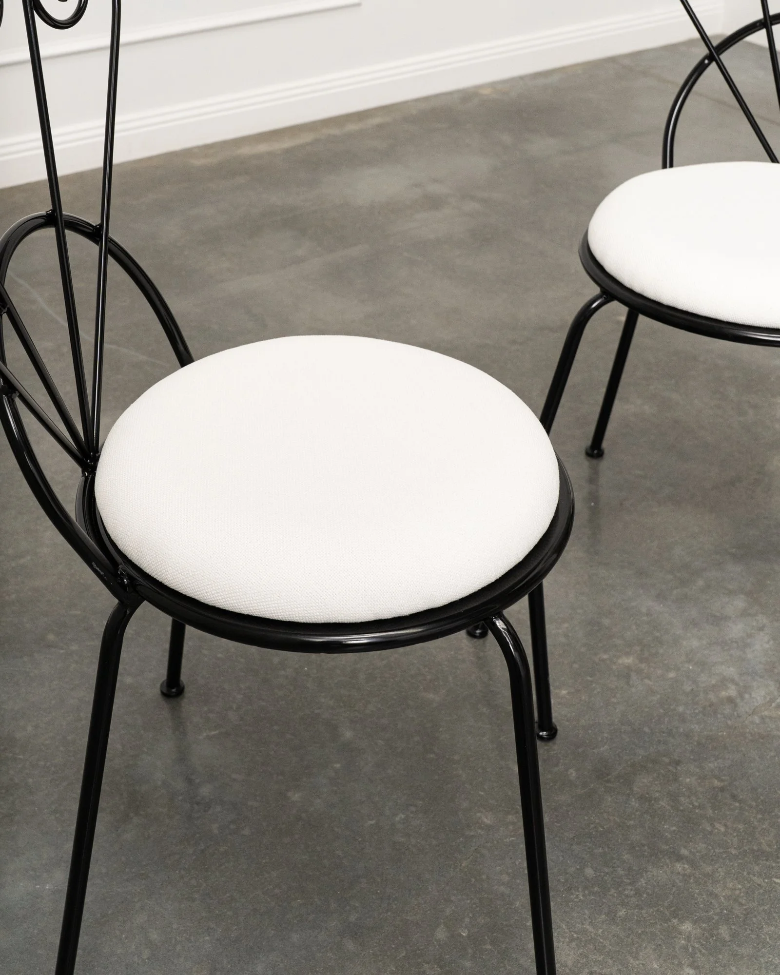 Two black metal framed chairs with white cushioned seats on a gray tiled floor, against a white wall and baseboards.