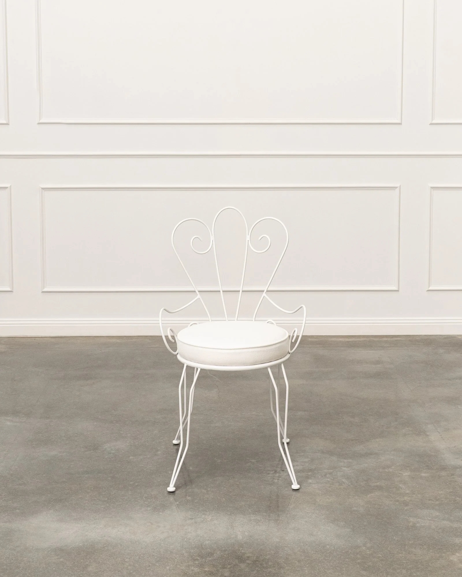 White decorative metal chair with heart-shaped backrest in front of a plain white wall with paneling.