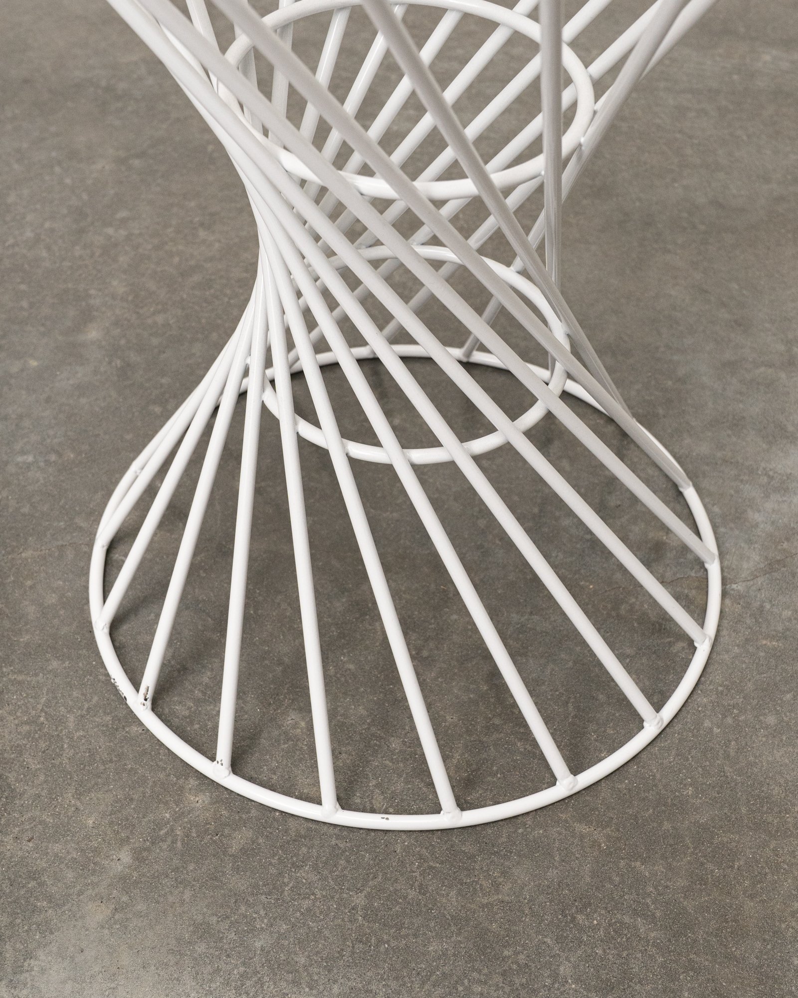 A white wireframe stool with a circular base and a rounded top, resting on a gray concrete floor.