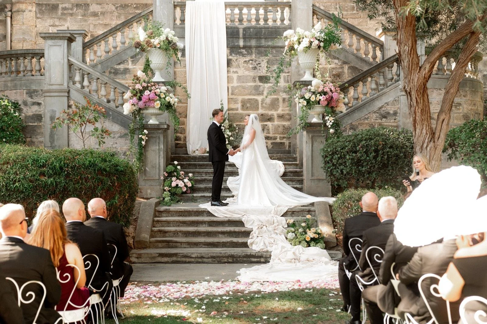 A wedding ceremony with a bride and groom holding hands on stone steps decorated with pink and white flowers, surrounded by guests seated on white chairs, with a woman officiant reading from a tablet on the right and floral arrangements on both sides of the steps.