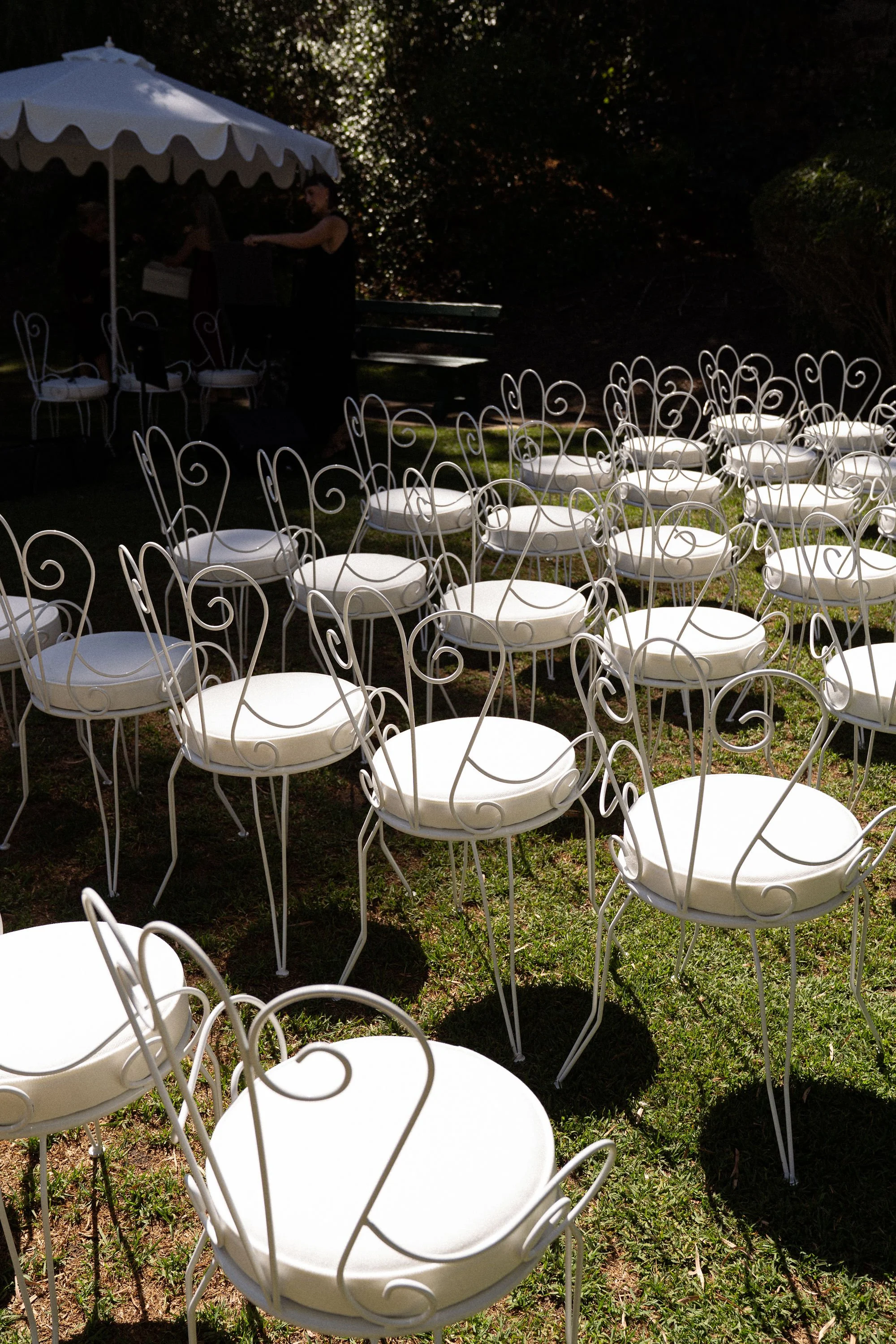 An outdoor setting with white metal chairs that have intricate, swirling design, arranged on the grass. In the background, there is a white canopy or tent and a few women gathering under it. The scene appears to be prepared for a gathering or event, possibly during evening or late afternoon.