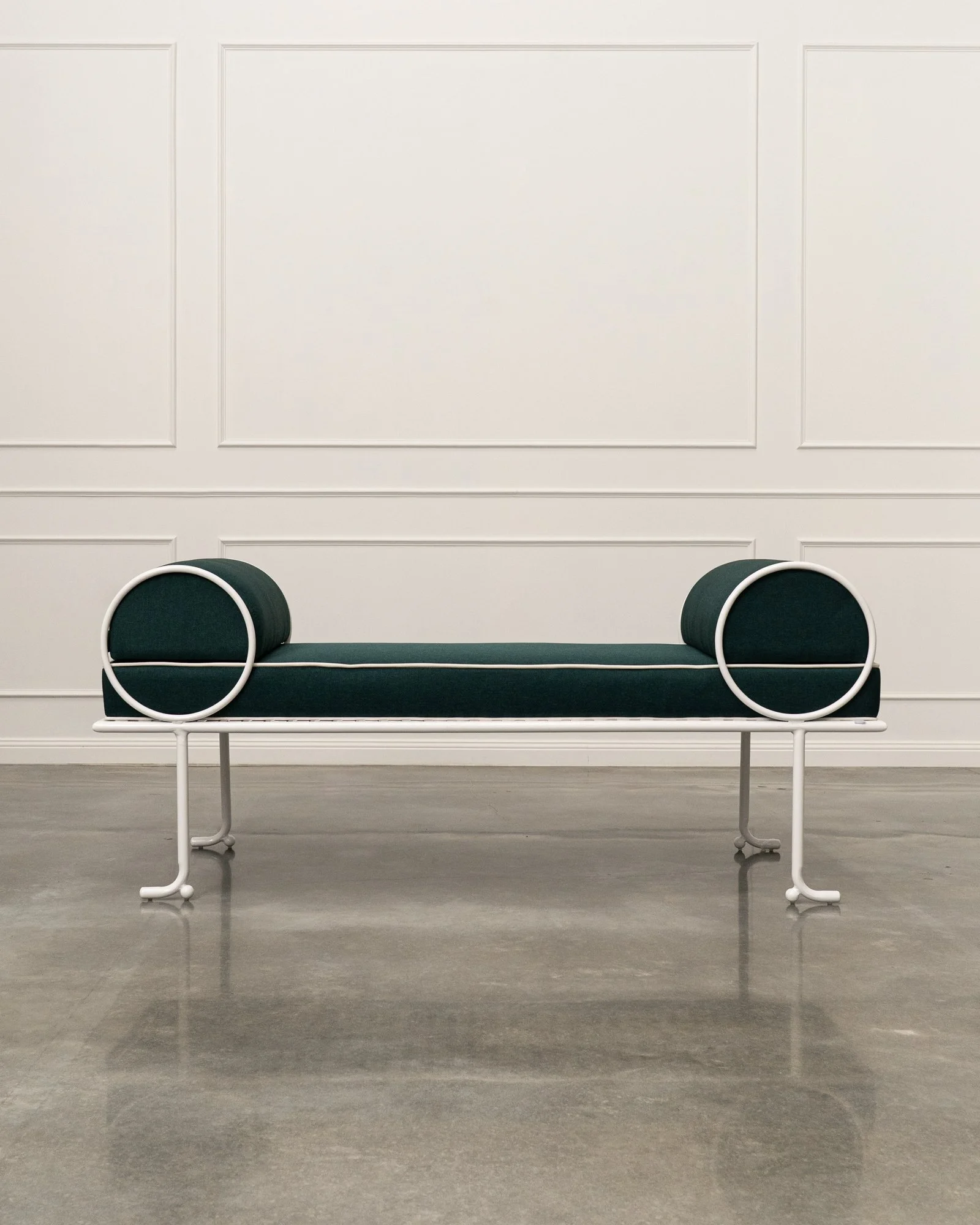 Minimalist modern bench with dark cushion and white metal frame, positioned against a plain white wall with subtle molding details.