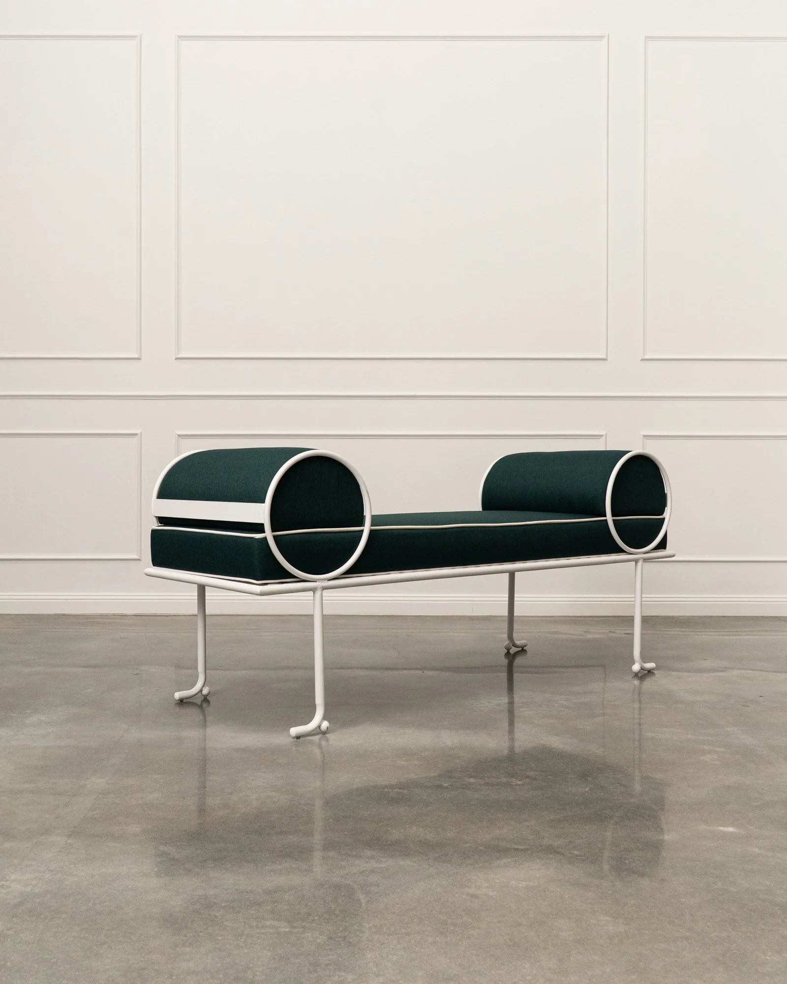 A modern bench with a black cushioned seat and backrest, white metal frame and armrests, situated in a minimalistic room with white wall panels and a grey concrete floor.