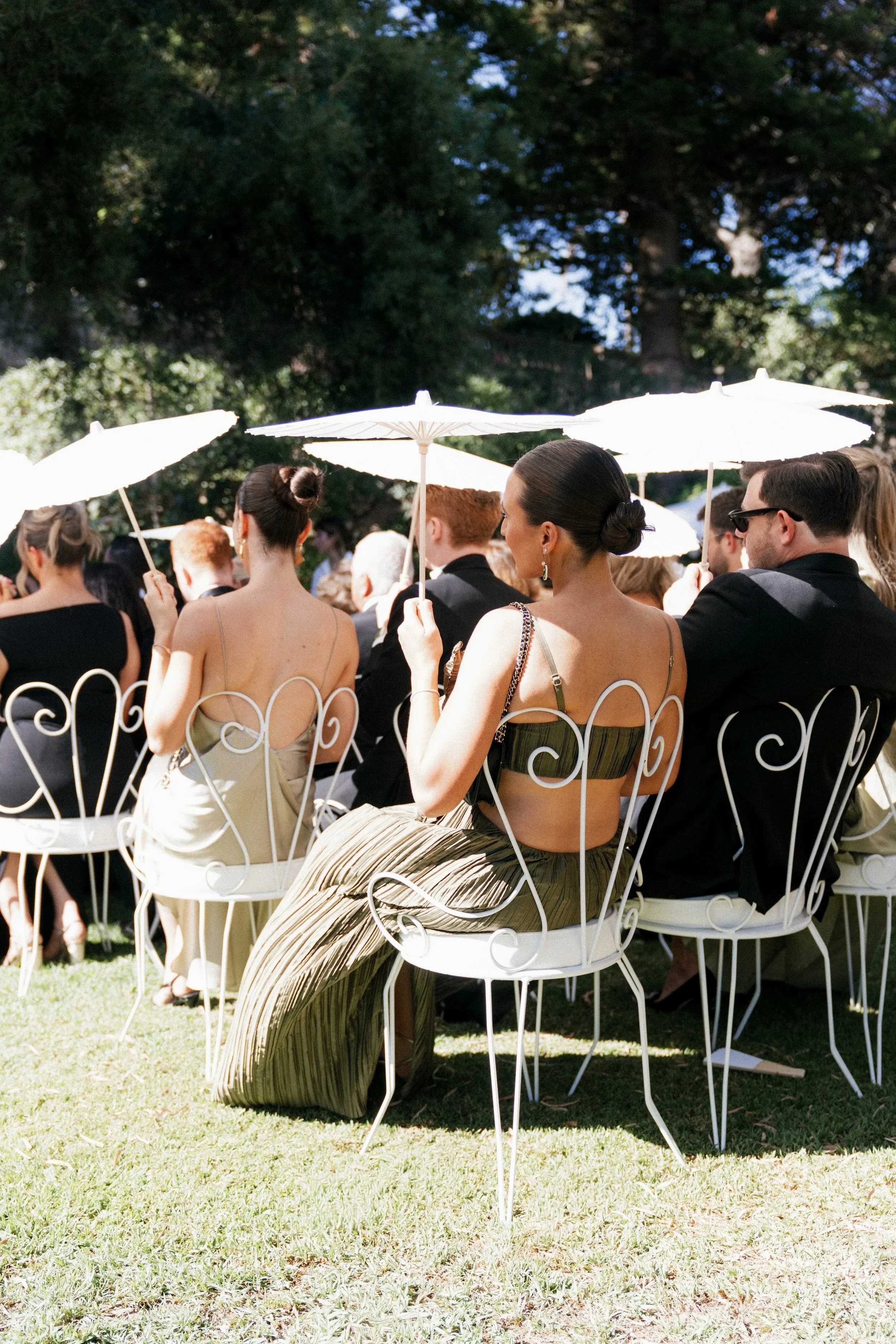 People attending an outdoor event, sitting on white metal chairs, holding white umbrellas, and dressed in formal attire, with trees and a grassy area in the background.