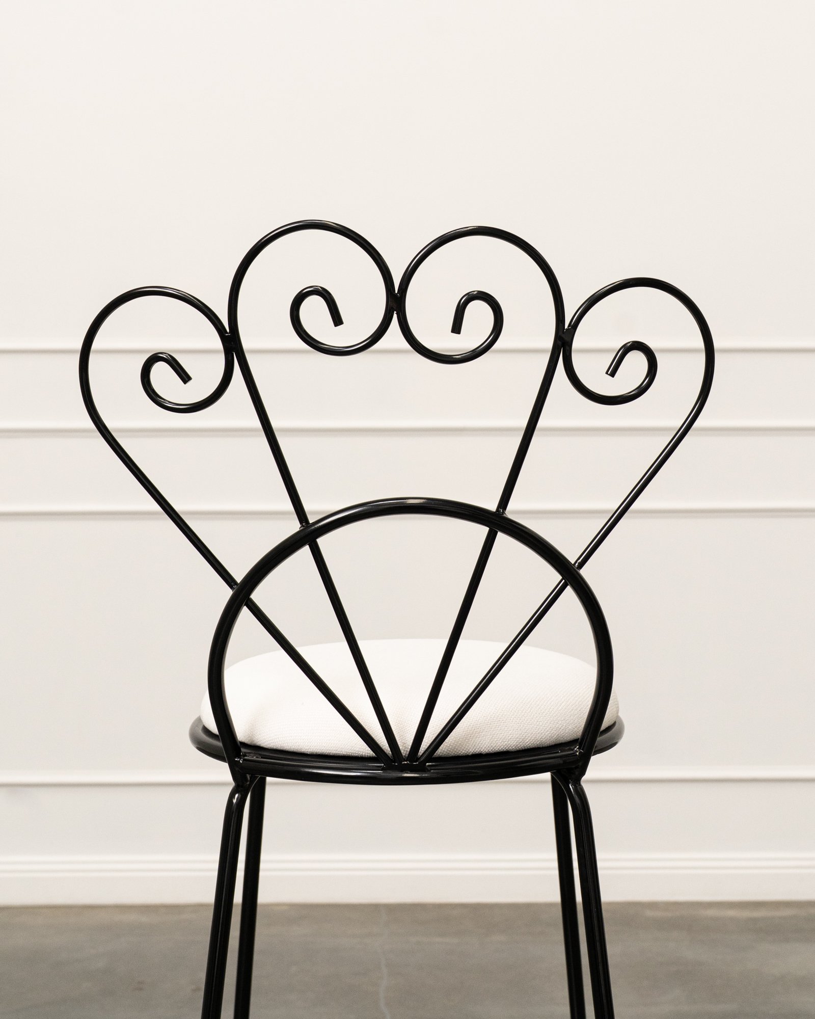 A black, metal, decorative garden chair with a padded white seat cushion, positioned against a white wall with horizontal paneling.