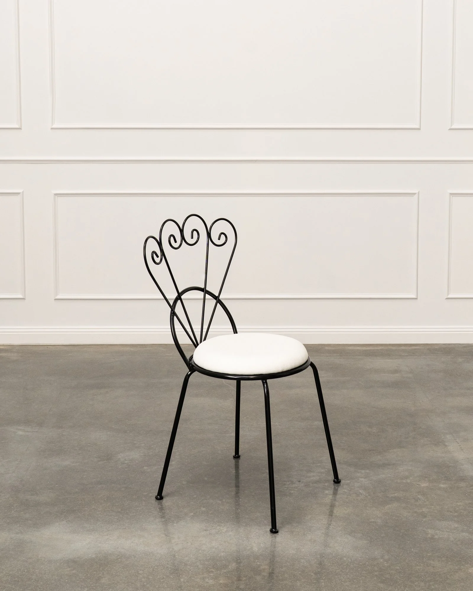 A black metal chair with a white cushioned seat against a white paneled wall on a gray floor.