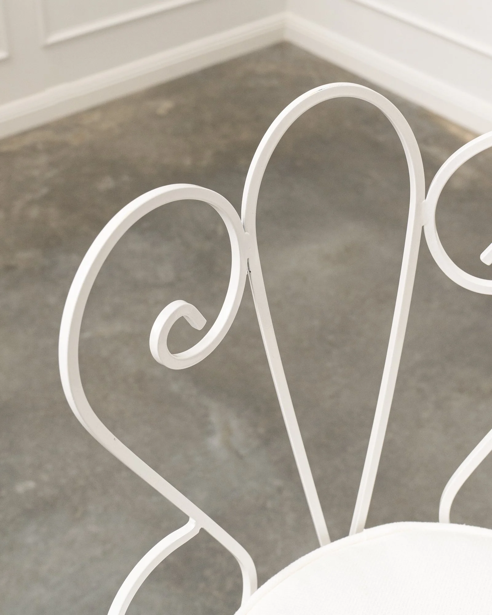Close-up of a white metal chair with decorative swirls in a room with gray flooring and white walls.