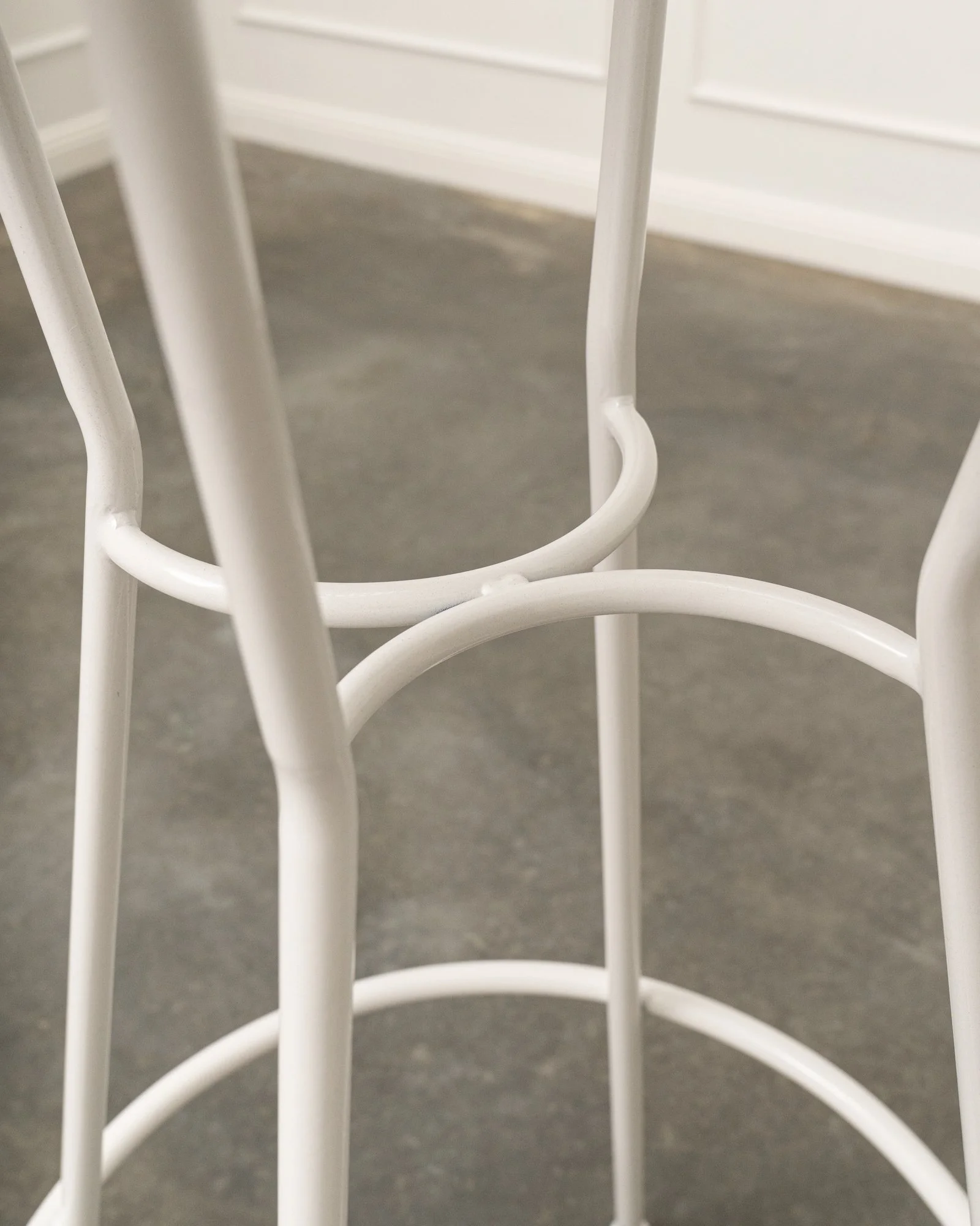 Close-up photo of white metal legs of a chair or table, with a gray floor and white wall in the background.