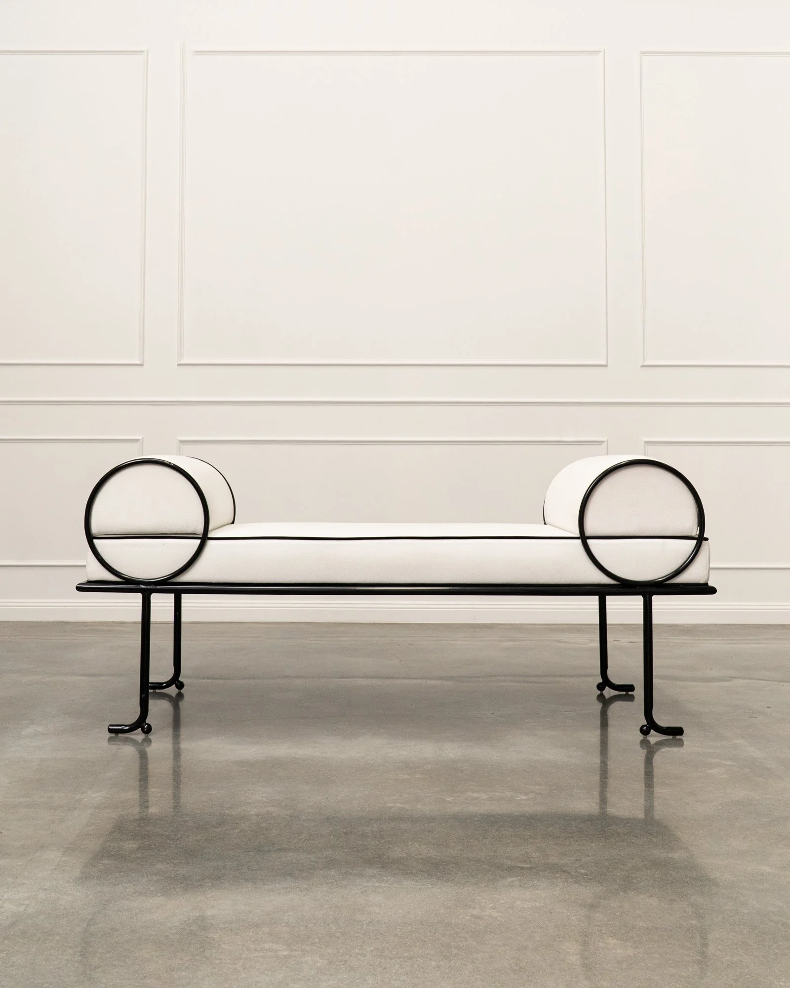 Minimalist modern bench with black metal frame and white cushioned seat in a bright, white-paneled room with a polished concrete floor.
