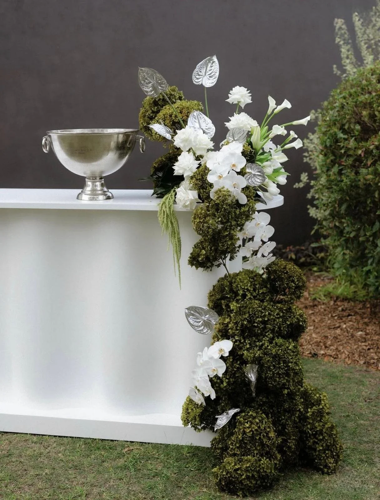 Outdoor scene with a white podium decorated with white flowers and greenery, and a silver bowl on top, against a dark background and outdoor greenery.