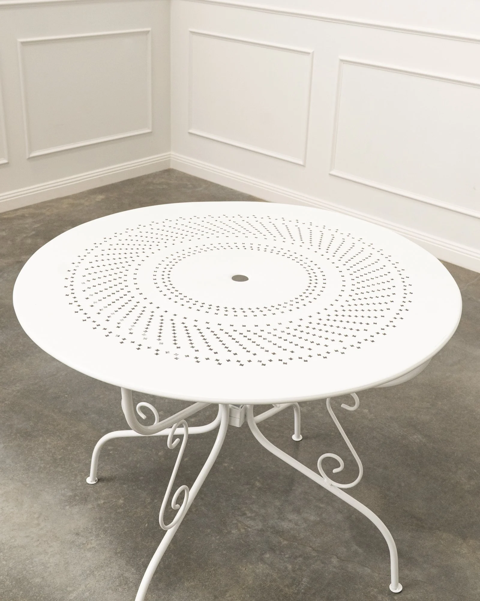 White round table with decorative metal legs and a perforated top design, placed on a dark gray floor against a white paneled wall.