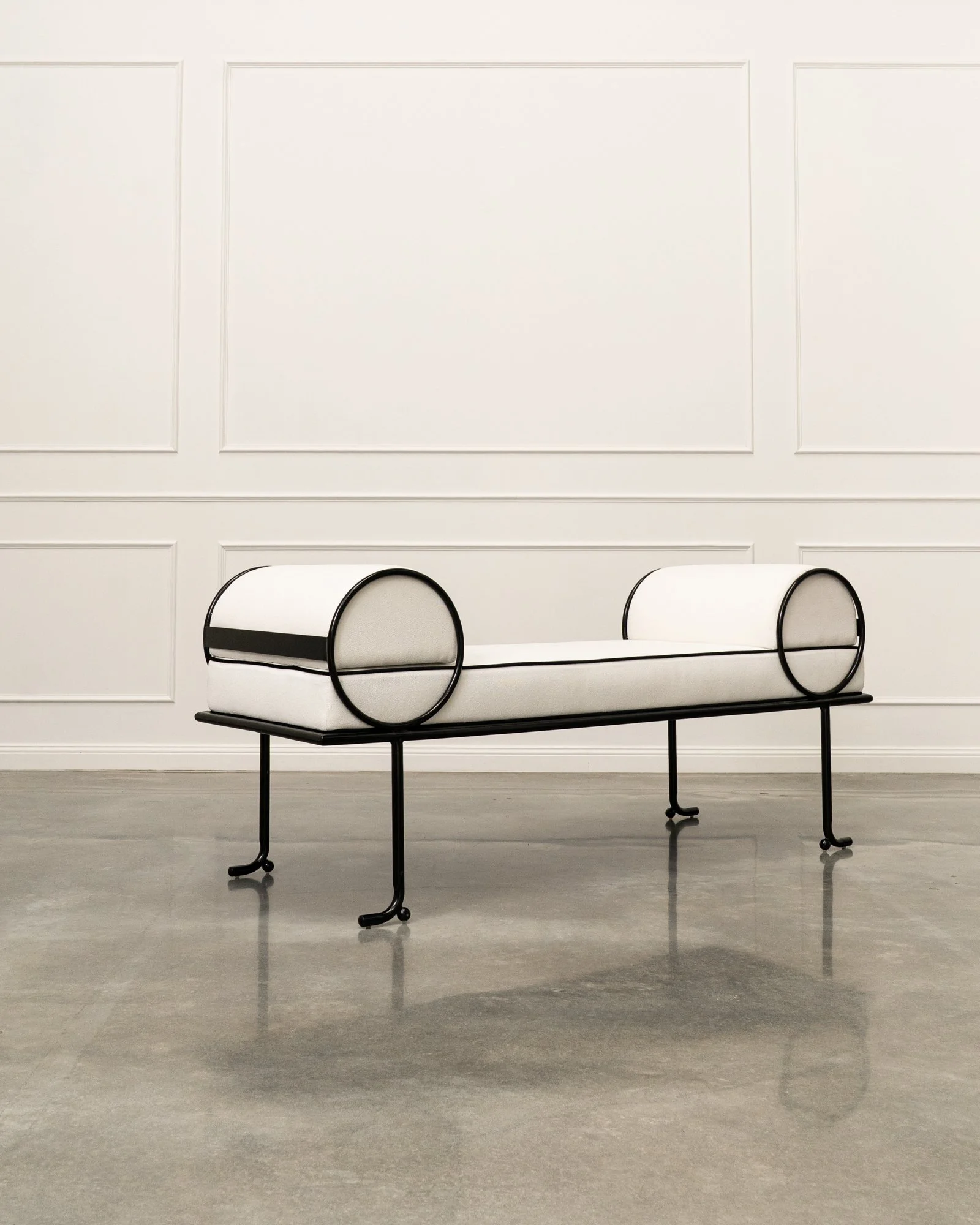Modern white bench with black metal frame and round supports, positioned against a white paneled wall in a gallery or showroom.
