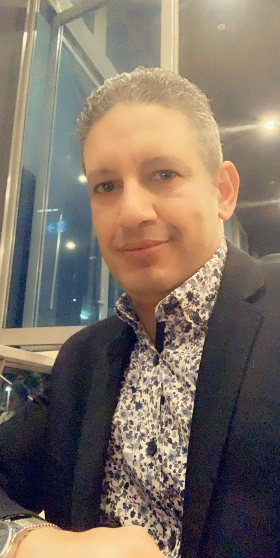 A smiling man with grayish hair wearing a black blazer and a patterned shirt sitting at a table in a restaurant or cafe with large windows and city lights outside.