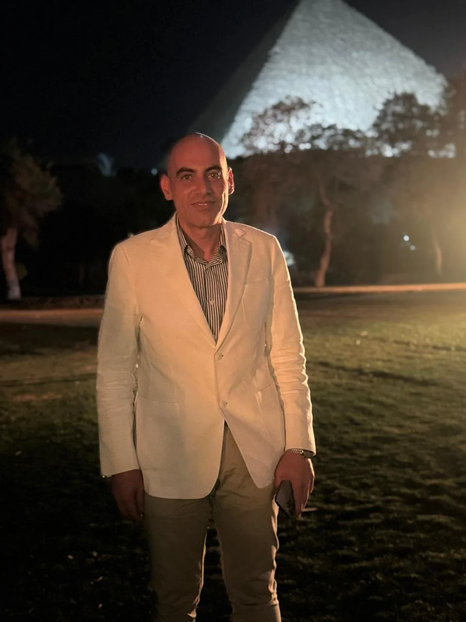 A man in a white jacket and beige pants standing outdoors at night, with a large illuminated sculpture or structure in the background.