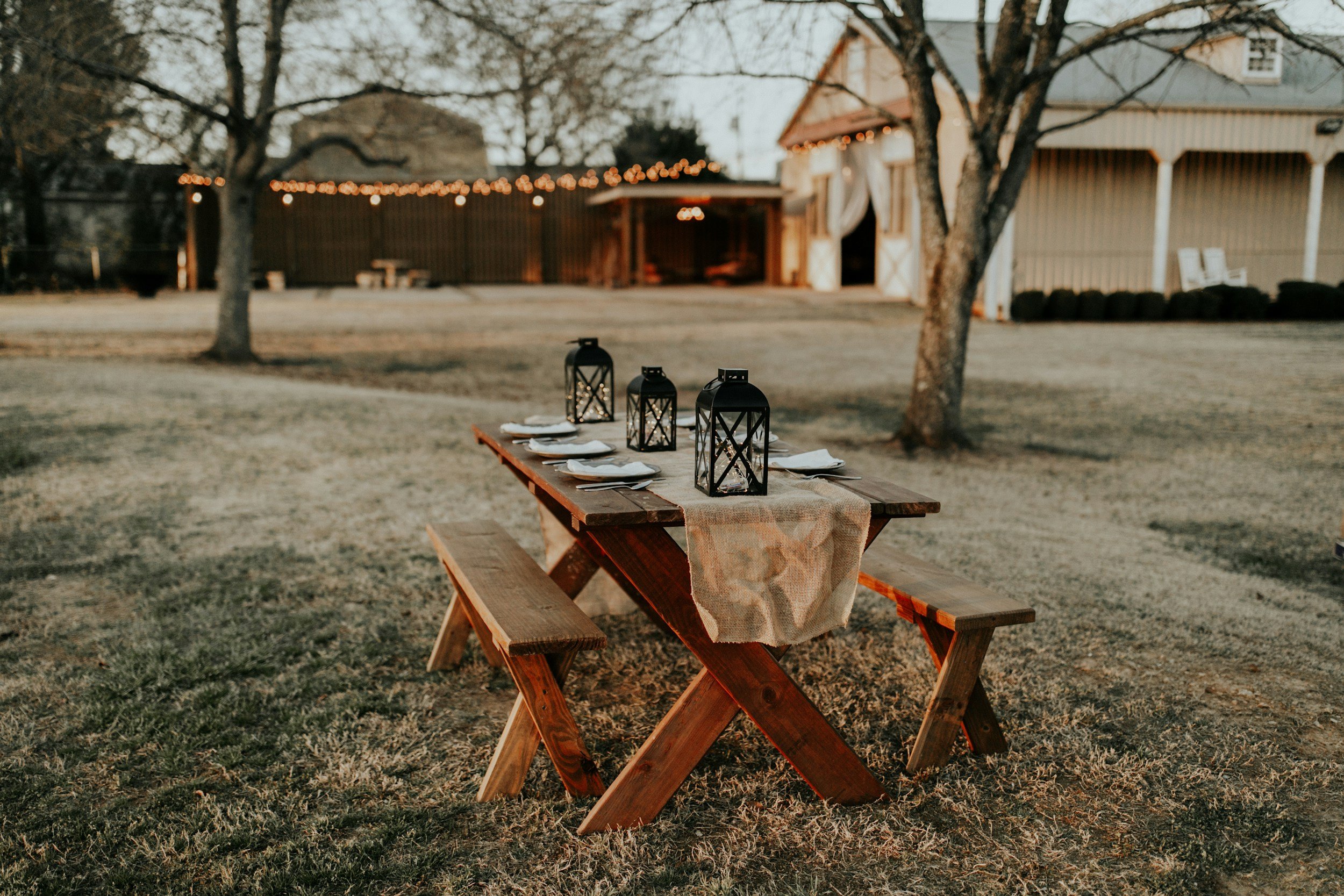 Outdoor dining setup with a wooden picnic table and benches on a grassy lawn, decorated with black lanterns and white plates, in front of a barn-style building with string lights at sunset.