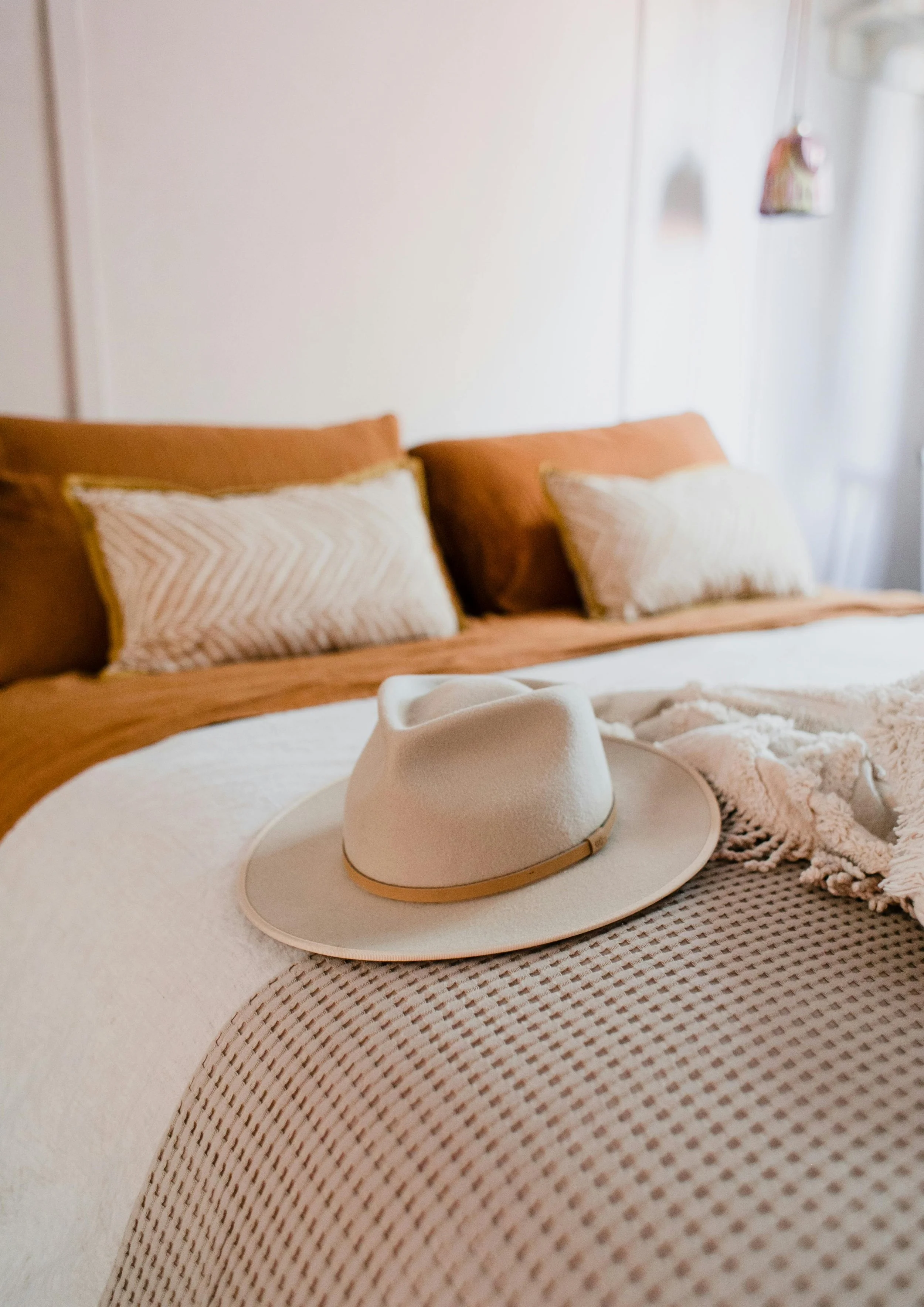 A white hat with a gold band resting on a round tray on a bed with beige and brown bedding, in a cozy bedroom with pillows and a window with curtains.