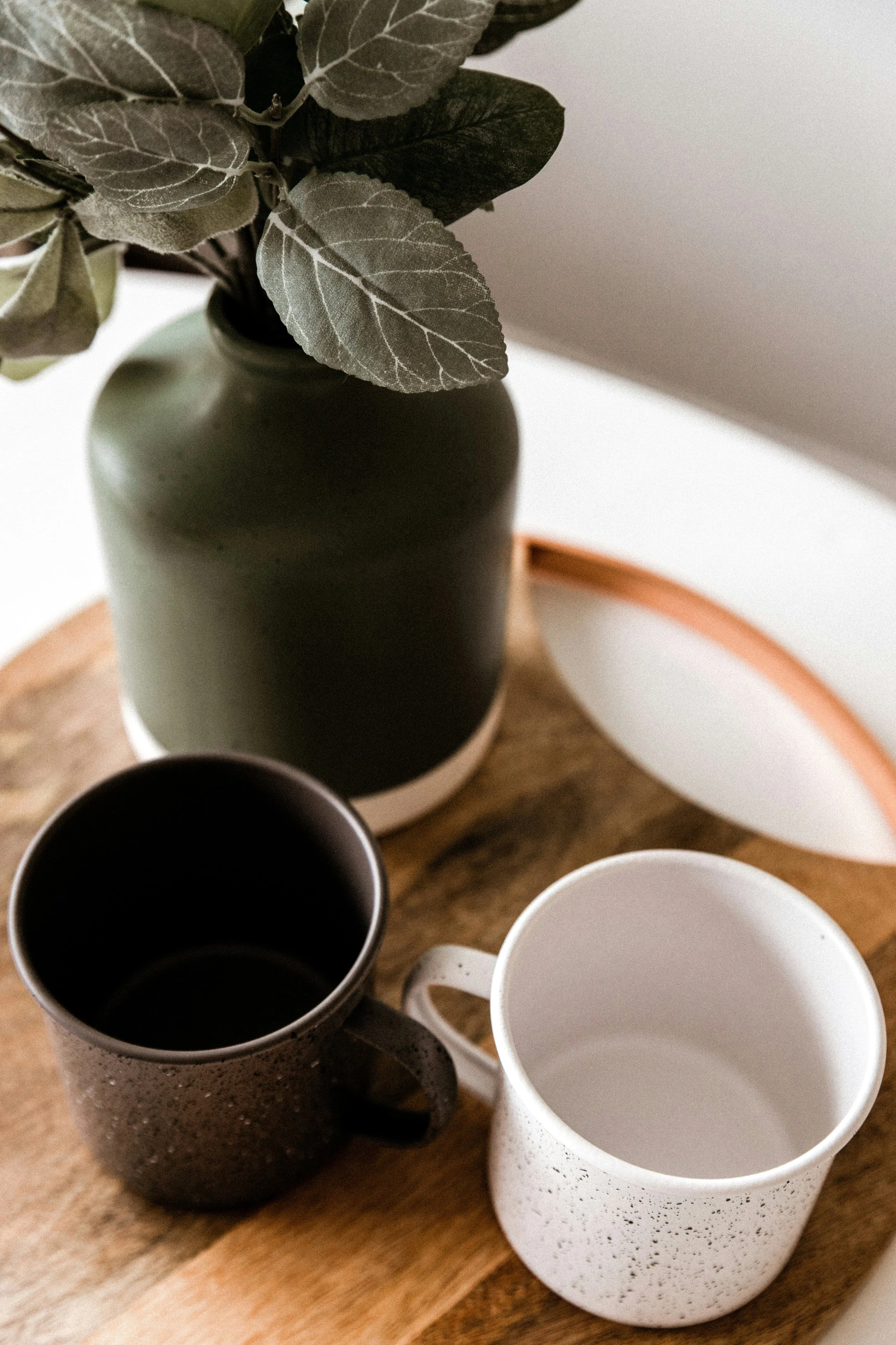 A dark green vase with a plant, two speckled mugs (one black, one white), on a wooden tray.