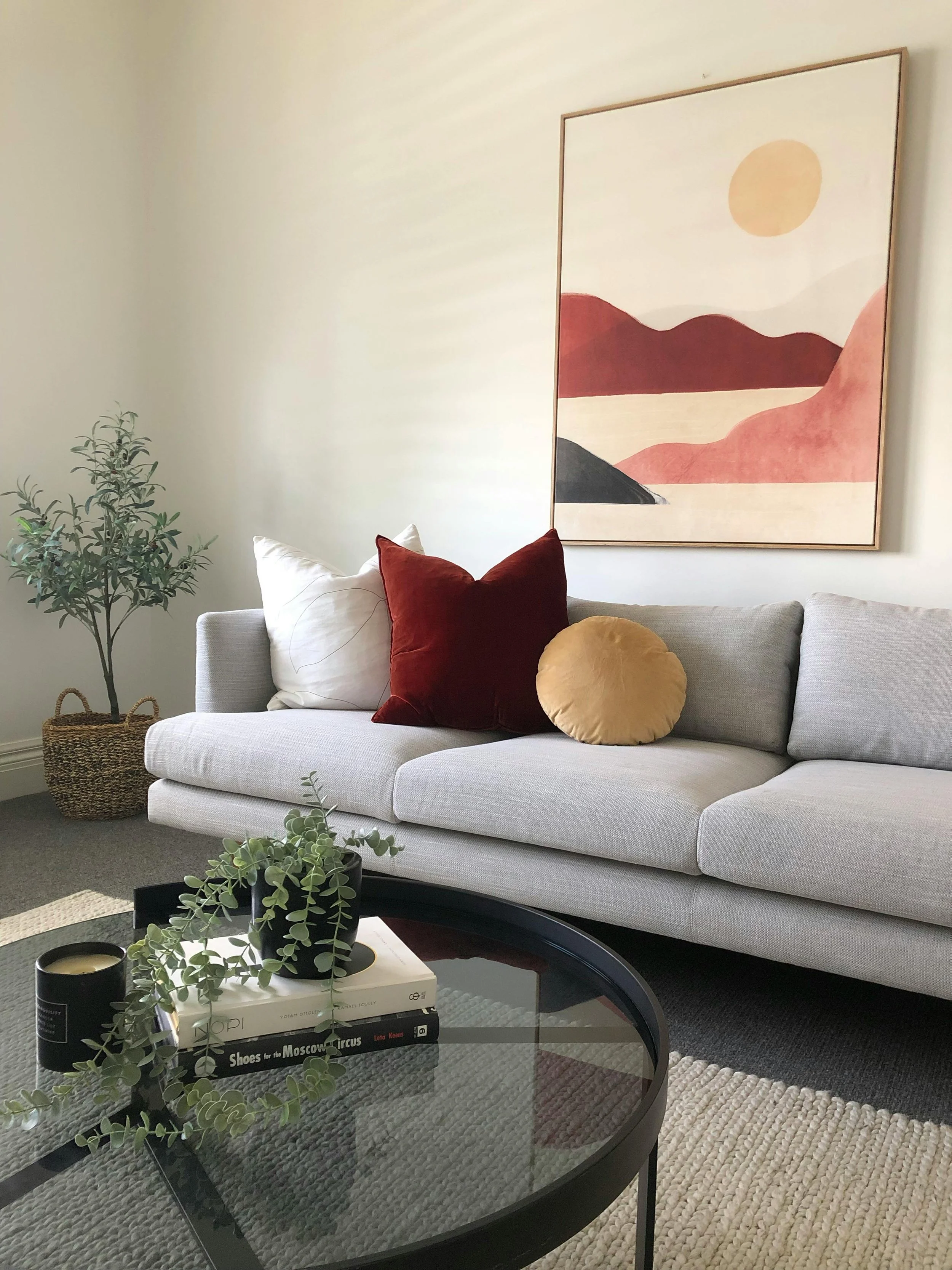 Living room with a light gray sofa decorated with white, red, and yellow throw pillows. A framed abstract landscape painting hangs above the sofa. There is a potted tree in a woven basket in the corner and a glass coffee table with books, a potted pl