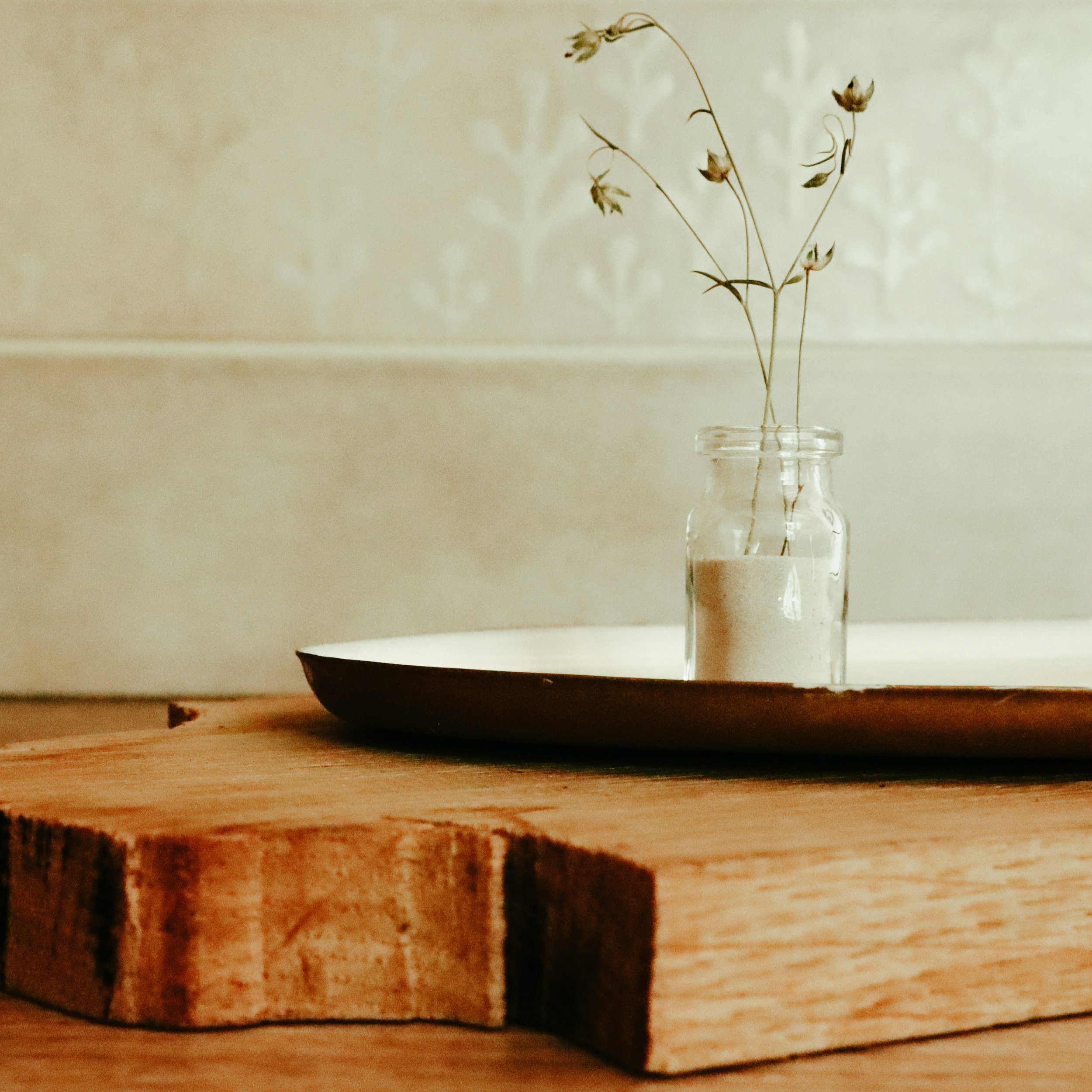 A small glass jar with dried flowers sits on a wooden board, which rests on a round tray against a patterned wall background.