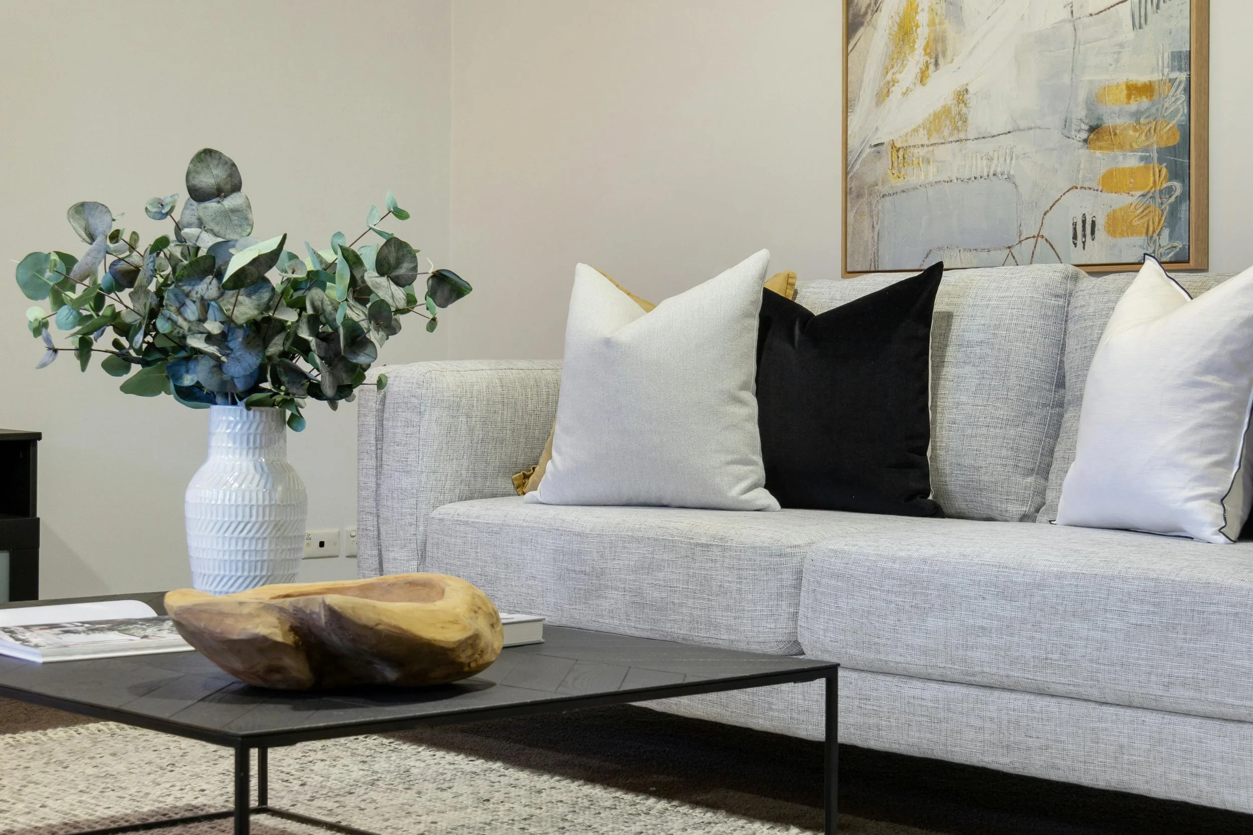 Gray living room sofa with white, black, and beige pillows, a black coffee table with a wooden decorative bowl and magazines, a white vase with green eucalyptus leaves, a textured rug, and an abstract paintings on the wall.