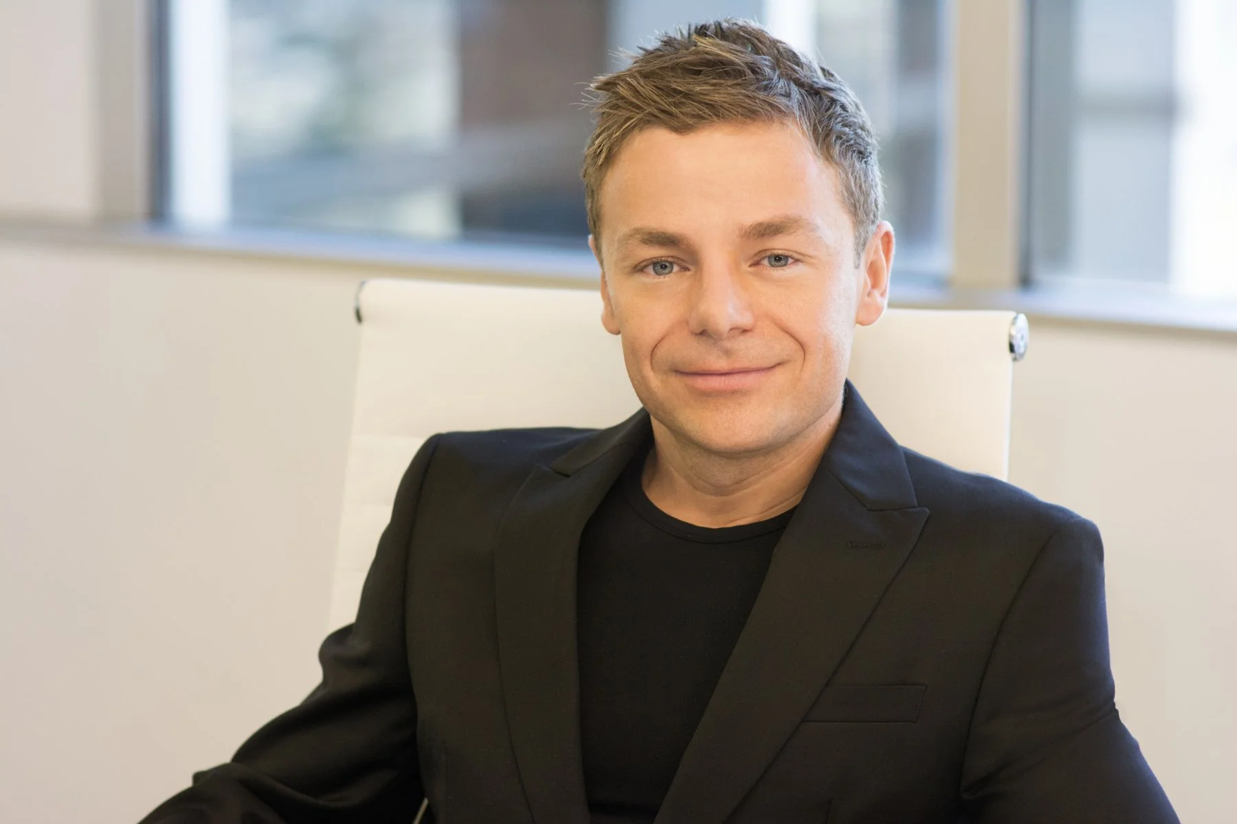 A young man with short brown hair and blue eyes, wearing a black blazer and t-shirt, is smiling and sitting in an office chair near a window.