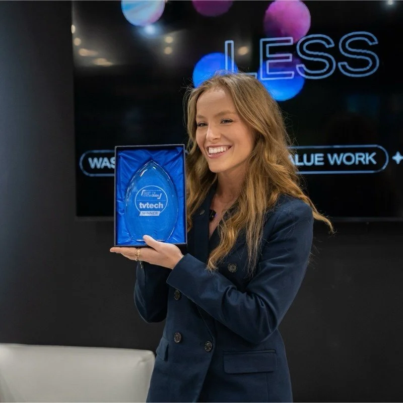 A woman with long red hair holding a clear glass award inside a blue box, smiling at an award ceremony. Behind her, there is a black screen with the word 'LESS' and other text, with colorful lights above.