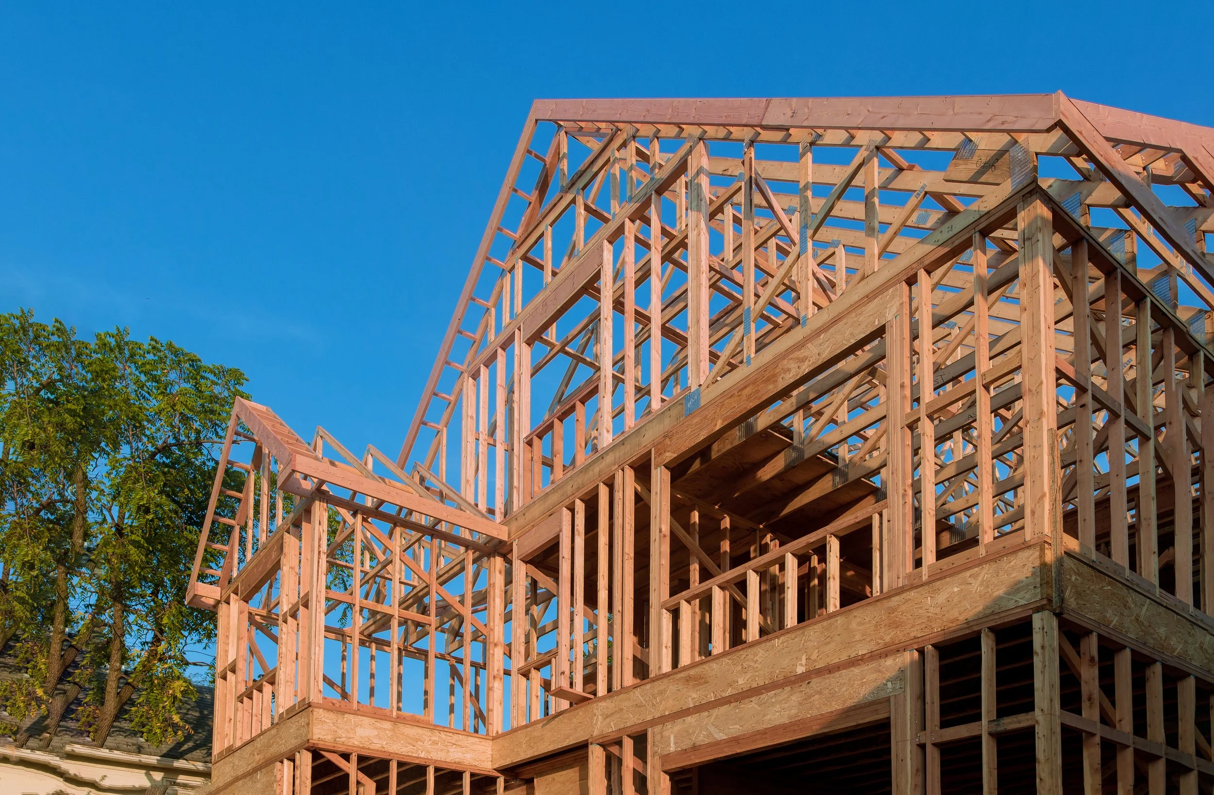 Wooden framing structure of a house under construction against a clear blue sky with a few green trees.