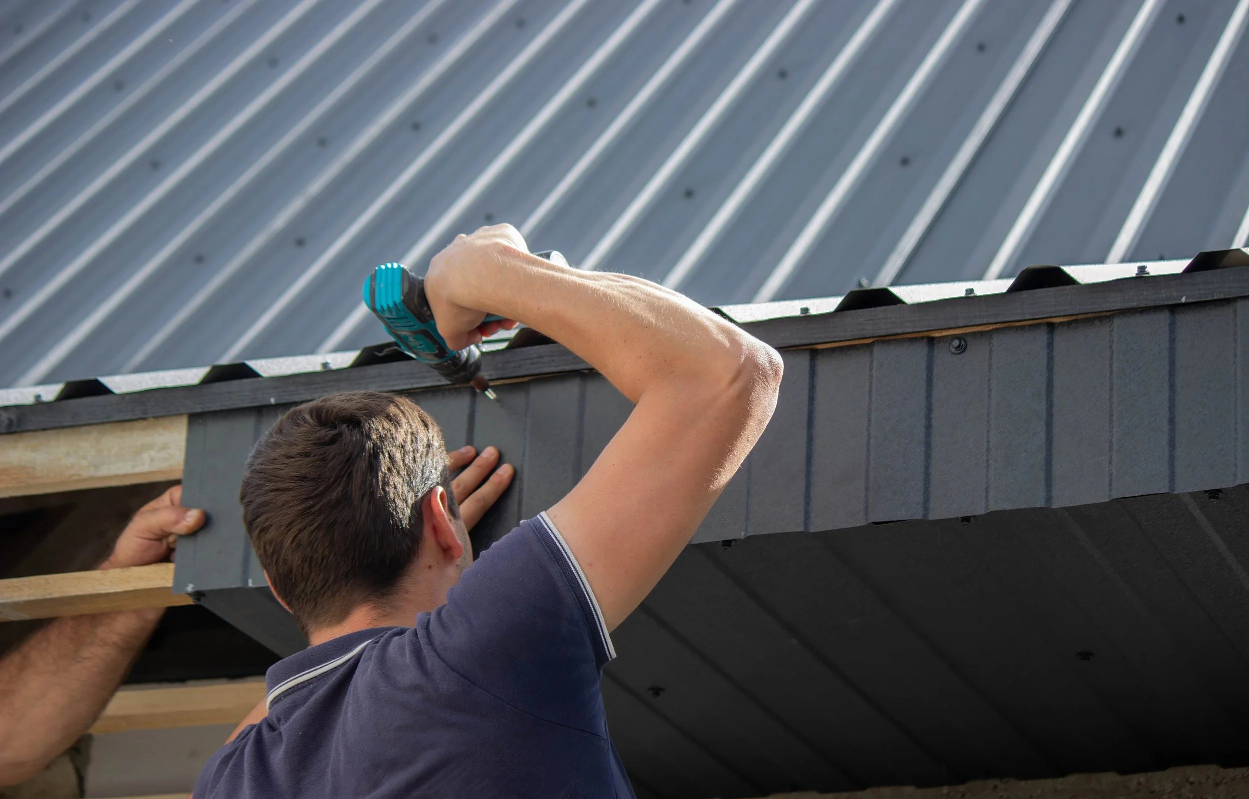 A person installing or repairing roofing on a building using a power drill, with the roof's edge and metal roofing panels visible.