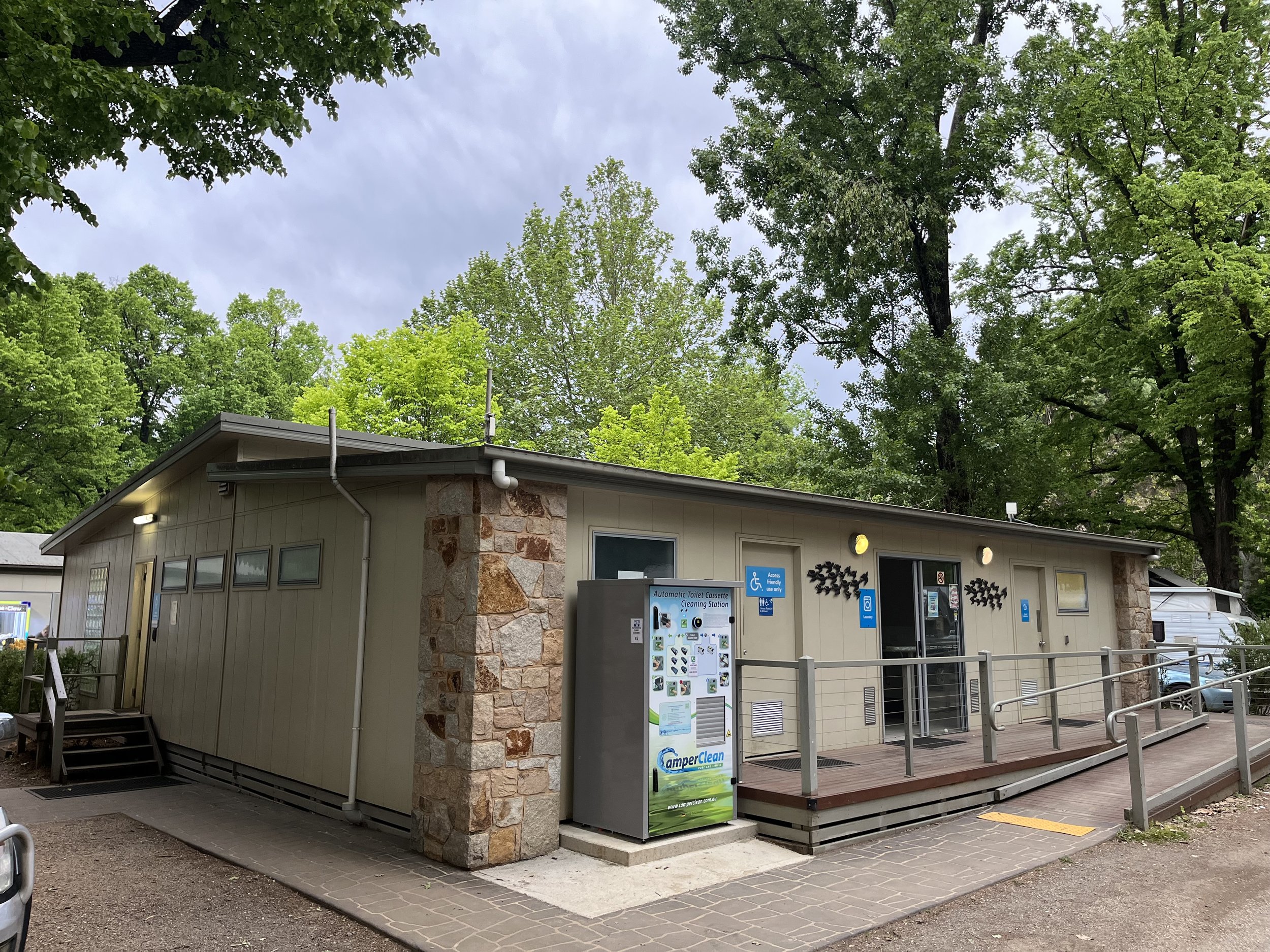 Public restroom building with a ramp, stone accents, several signs indicating accessibility, and a vending machine outside, surrounded by green trees.