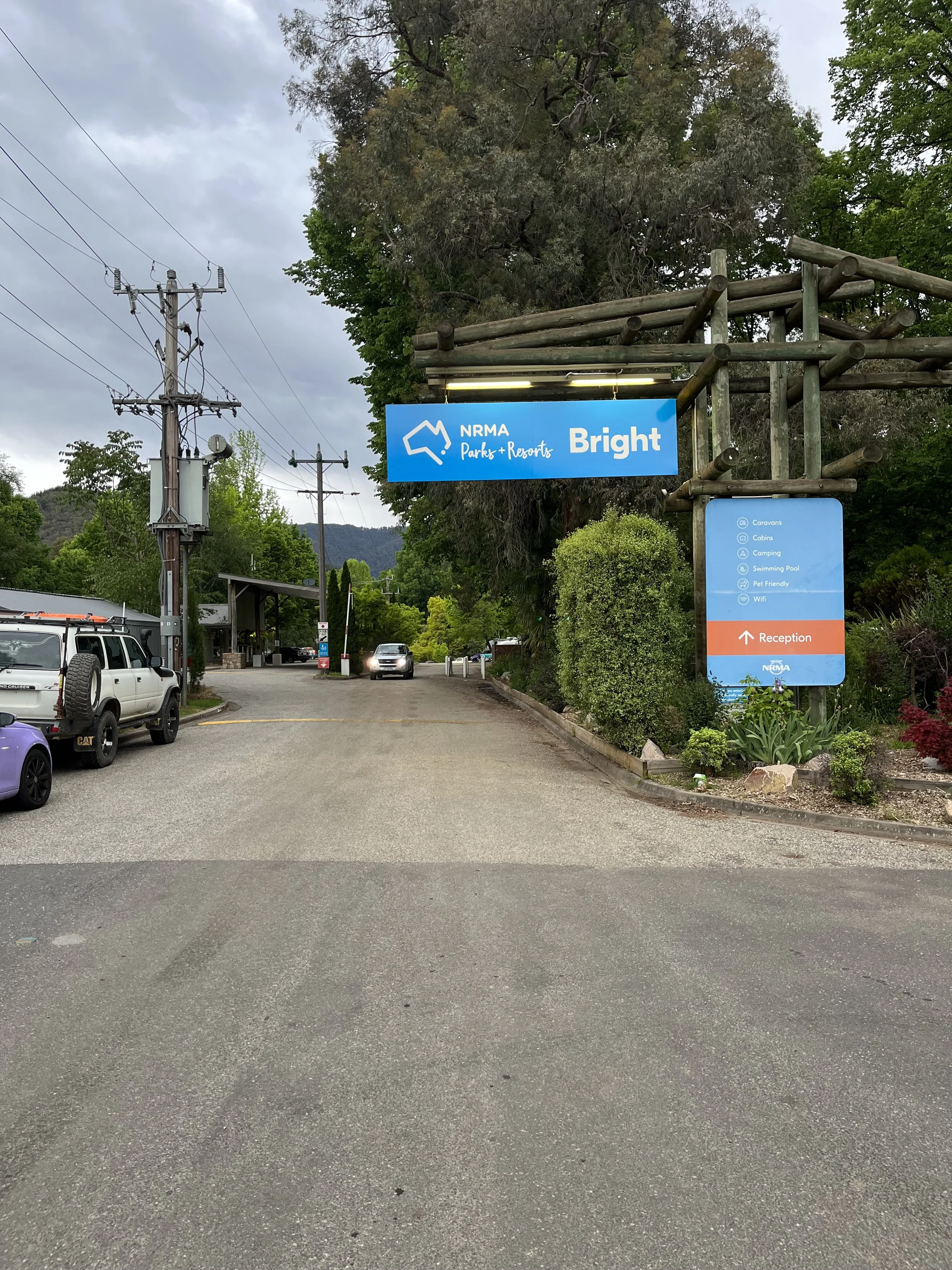 Entrance to NRMA Parks and Resorts Bright featuring a blue sign with the NRMA logo, listing amenities such as caravans, cabins, camping, swimming pool, pet friendly, and WiFi, with an arrow pointing to reception.