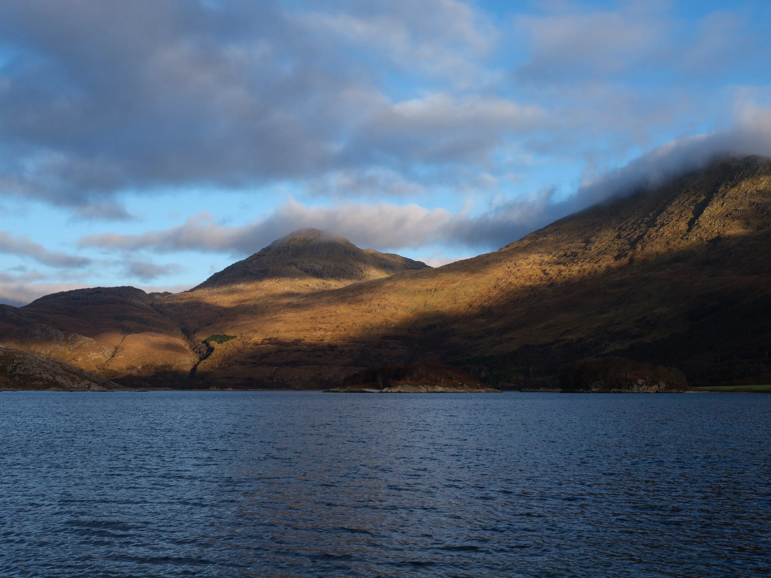 A landscape featuring a lake in the foreground and rolling hills and mountains in the background, with partly cloudy skies overhead.