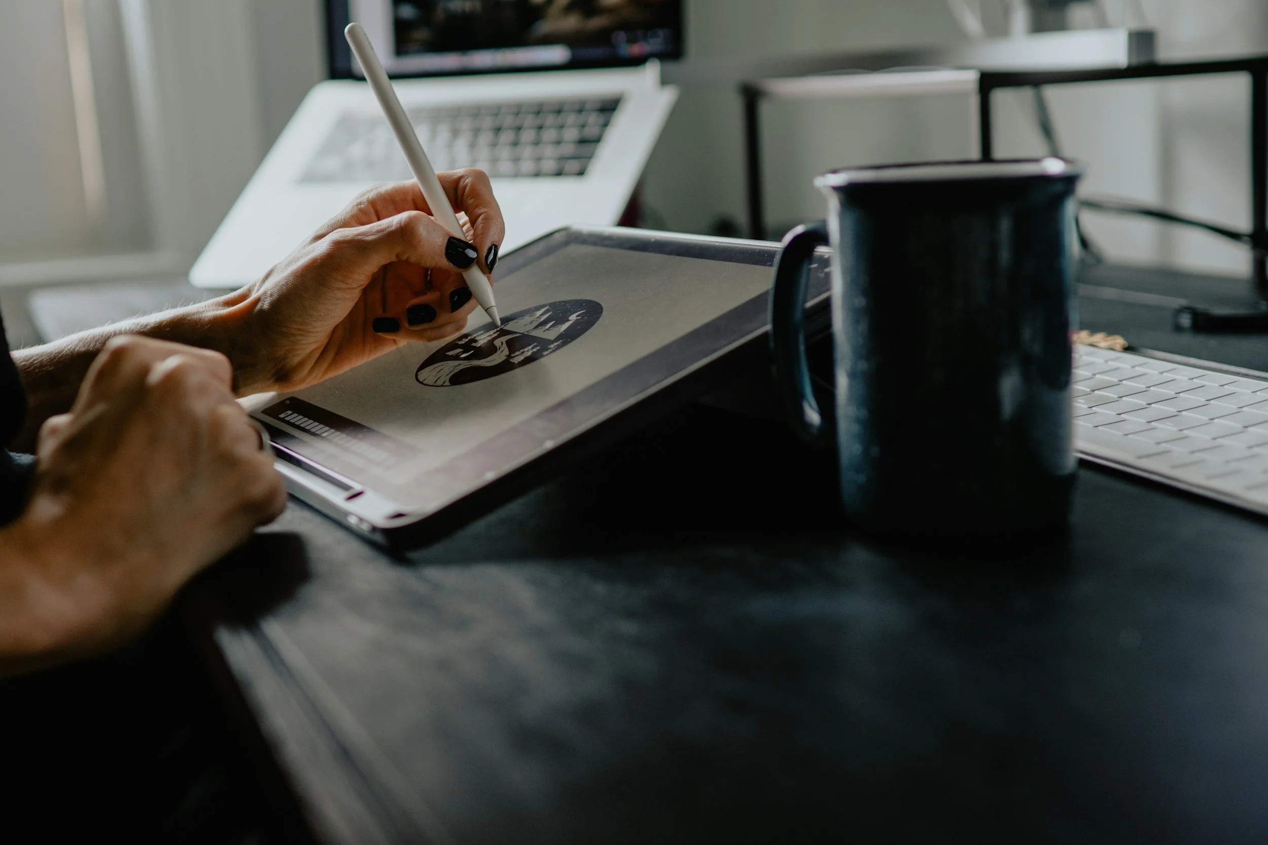 Person using a digital pencil on a drawing tablet, with a mug, keyboard, and laptops on a dark desk.