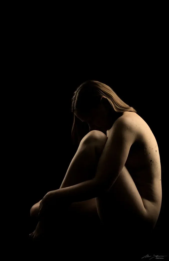 Nainen istuu polvet sylissä,  mustassa taustassa , valaistuna sivulta.