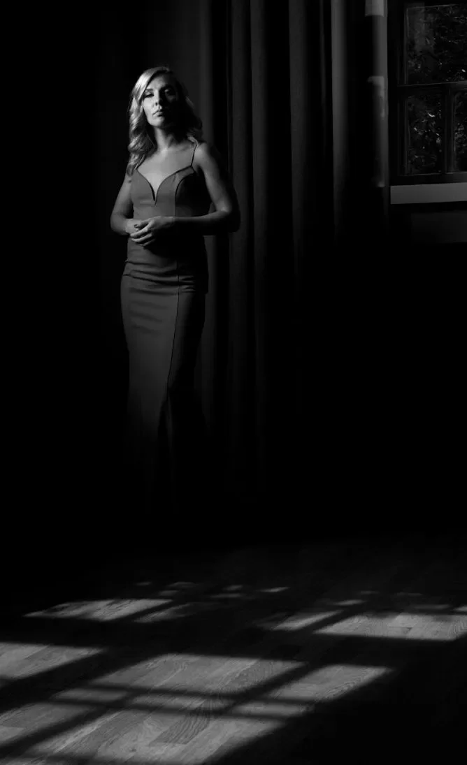 Tummasävyinen kuva, jossa nainen seisoo ikkunan vieressä pimeässä huoneessa, ikkuna heijastaa valoa ja varjoja lattialle.