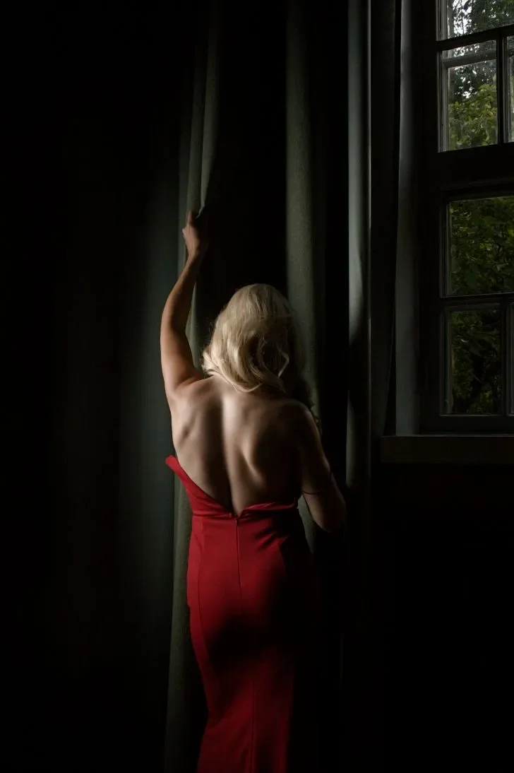 Nainen seisoo ikkunan äärellä, katsoo ulos, ja hänen selkänsä näkyy. Hänellä on punainen mekko ja hän nostaa kättään verhoa pitkin.