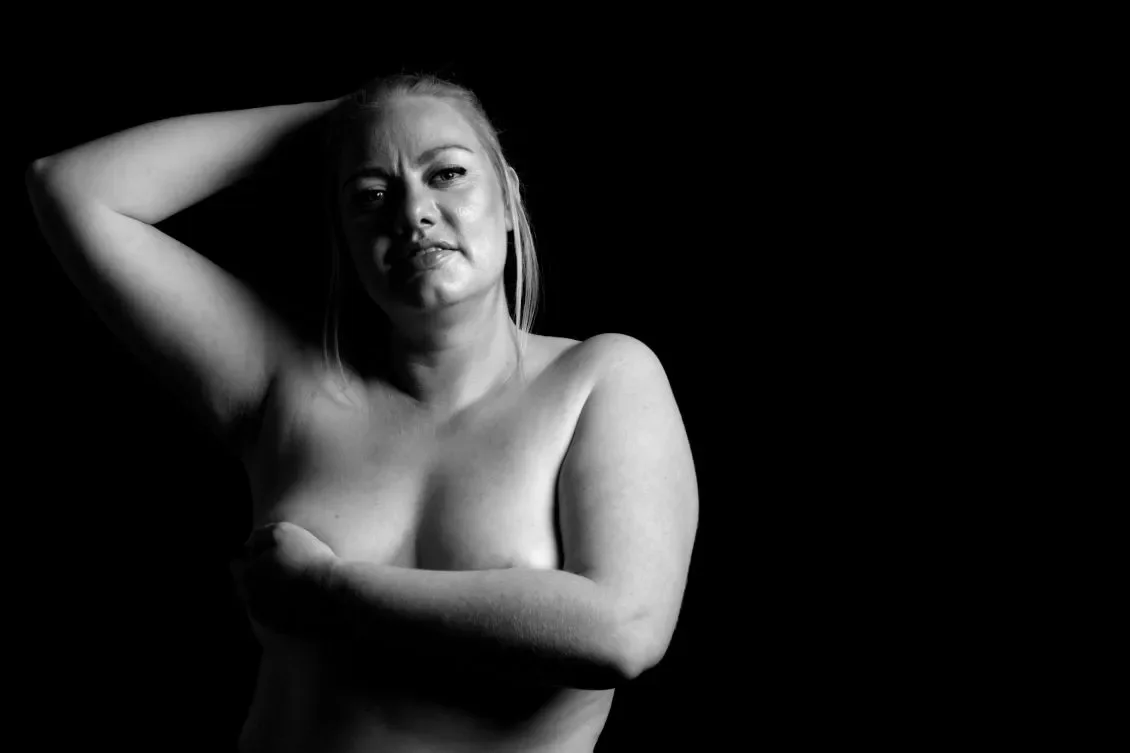 Mustavalkoinen kuva naisesta. Käsi rintojen päällä ja toinen käsi pään takana, katsoo suoraan kameraan.