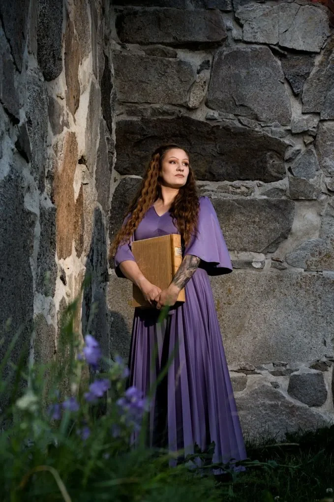 Nuori nainen, jolla on pitkät kiharat hiukset, seisoo kivisen rakennuksen seinän vieressä. Kädessä iso kirja.