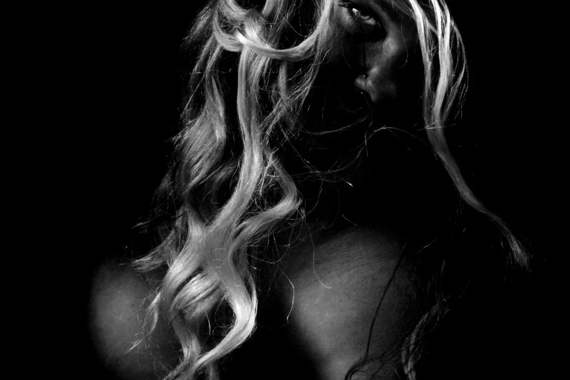 Tummasävyinen kuva naisesta jonka pitkä, kiharainen hius peittää osittain hänen kasvonsa.