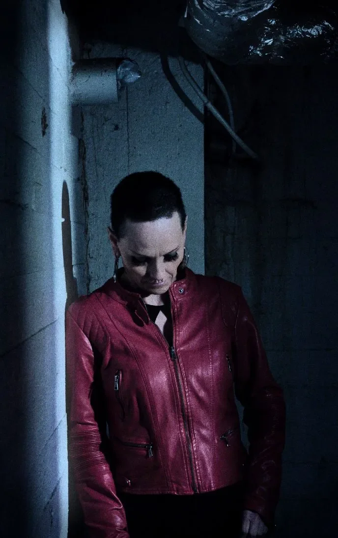 Ihminen punaisessa nahkatakissa seisoo seinän vieressä tummassa ympäristössä.