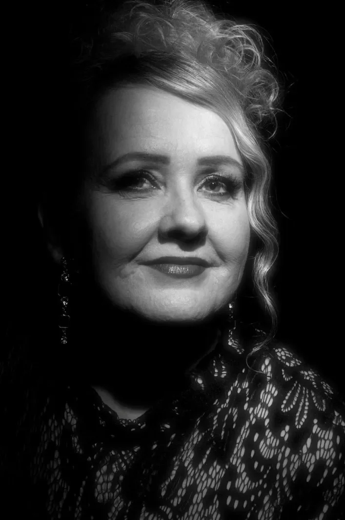 Mustavalkoinen valokuva naisesta, jonka hiukset ovat kiharat. Pieni hymy kasvoilla. Tumma kuva, mutta naisen kasvoille tulee valoa.