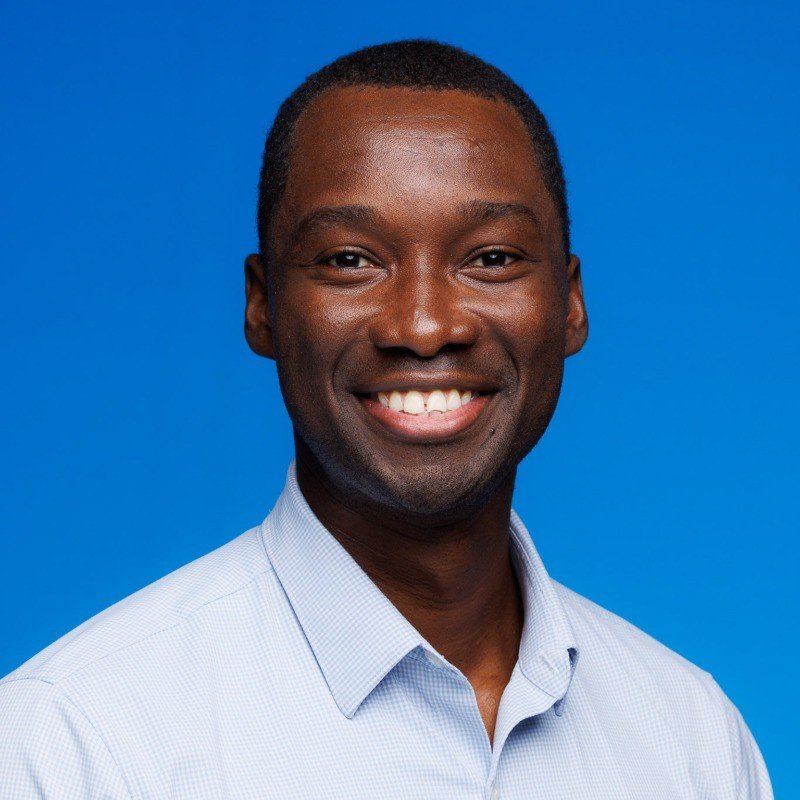 Portrait of a smiling Black man with short hair, wearing a light blue collared shirt against a blue background.