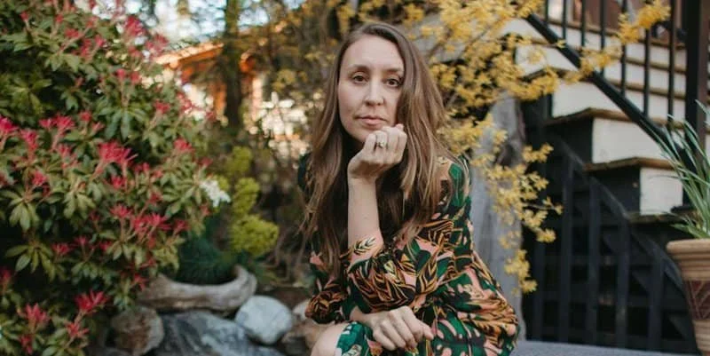 Woman with long brown hair sitting outdoors near colorful plants and a staircase.