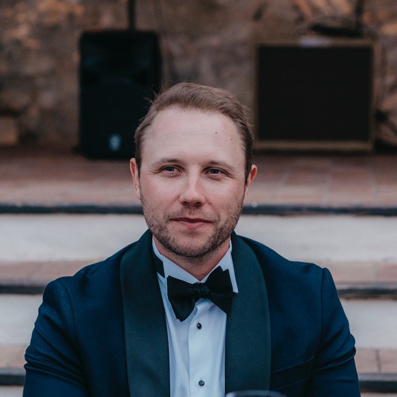 Portrait of a man in a tuxedo with a bow tie, sitting on stairs, with a brick wall and speakers in the background.
