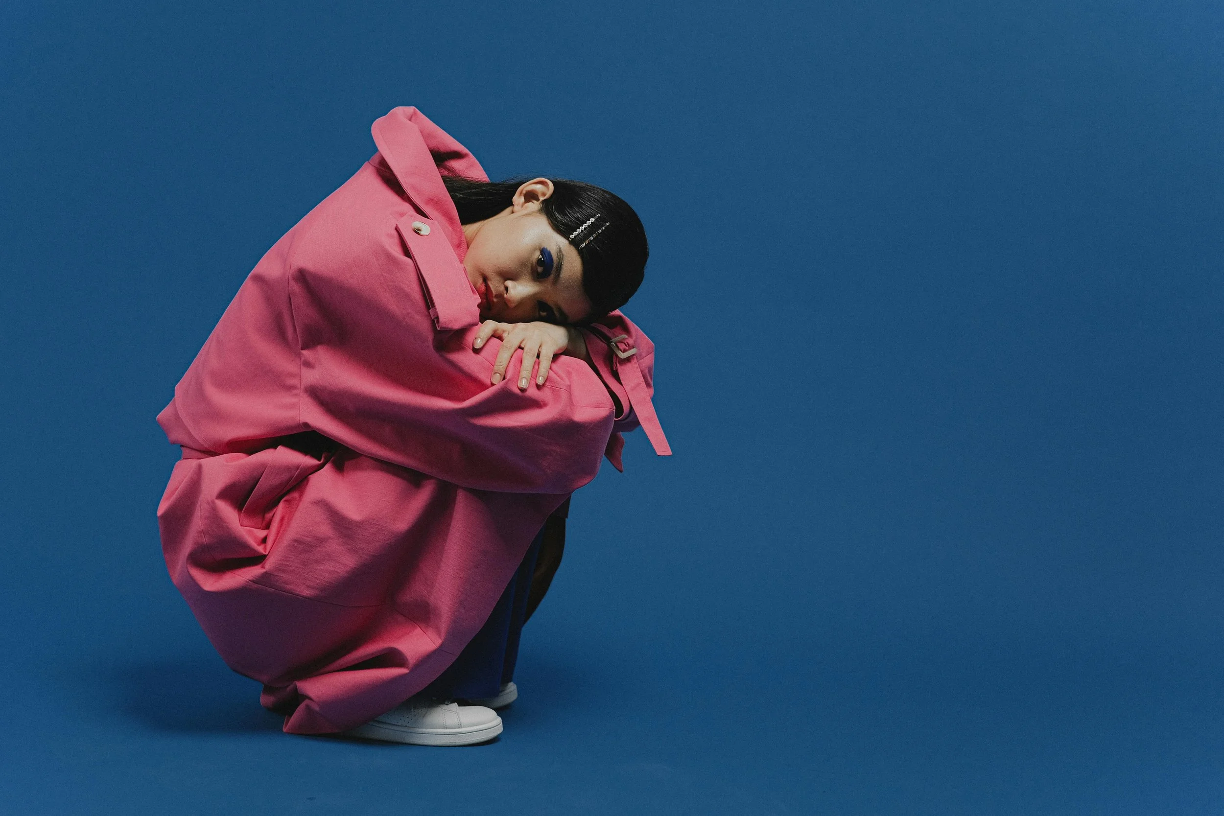 Young woman crouching against a blue background, wearing a pink oversized coat with her head resting on her knees, looking at the camera.