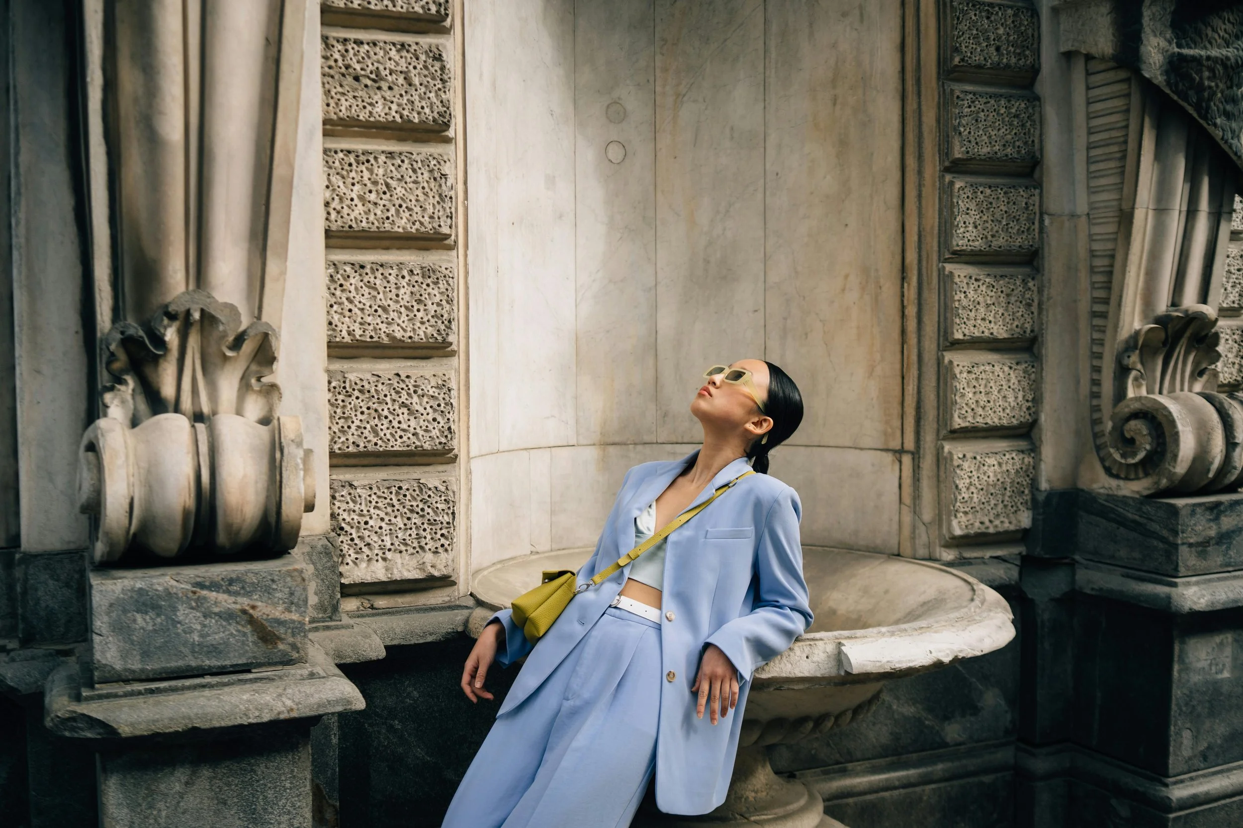 Woman wearing a light blue suit with a yellow bag and sunglasses leaning back in an ornate stone fountain surrounded by classical architecture.