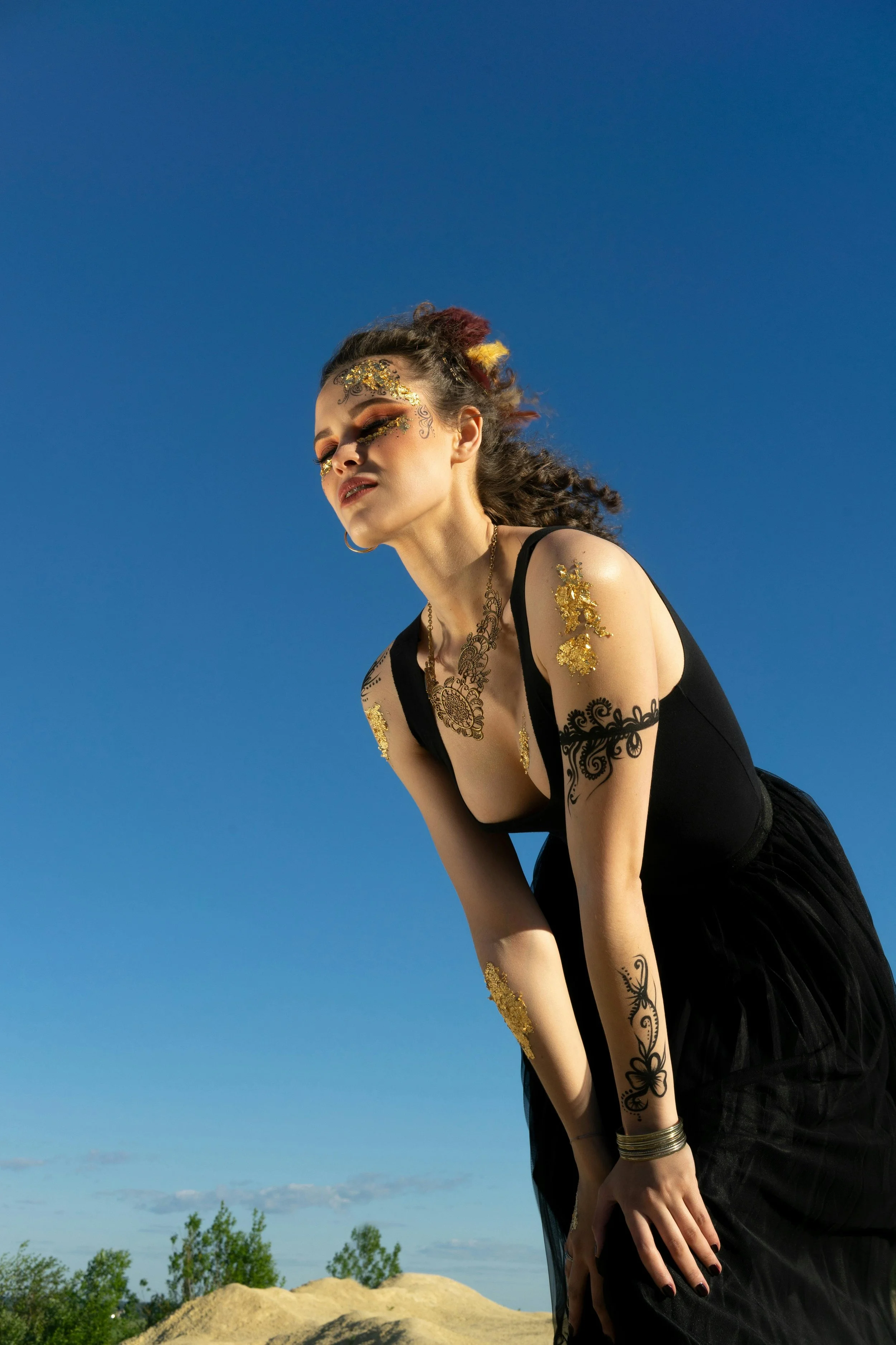 A woman with gold decorative makeup and tattoos, wearing a black dress, leaning forward outdoors against a clear blue sky.