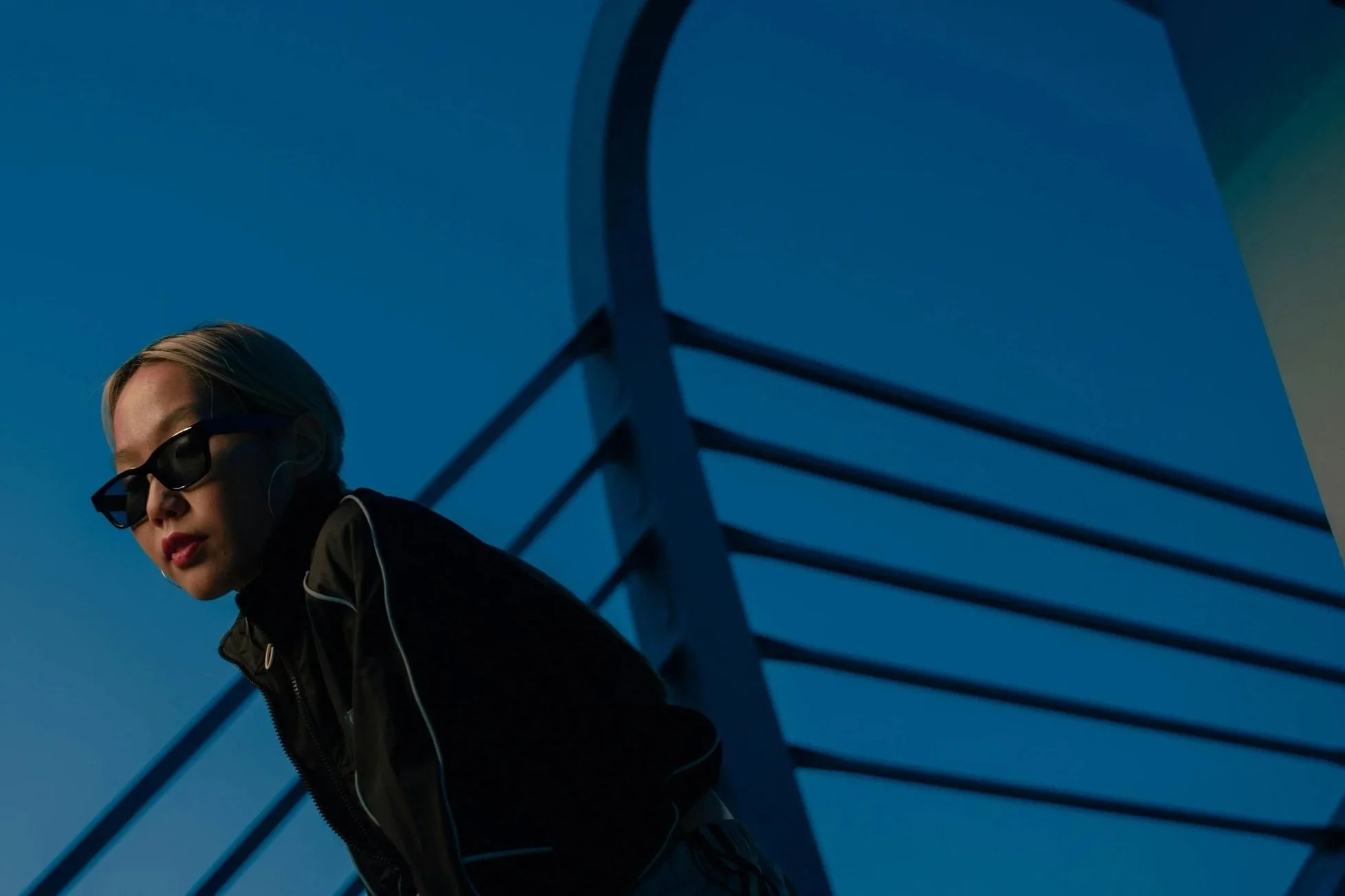 A woman with blonde hair wearing sunglasses and a black jacket, leaning against a modern architectural structure with a blue sky background.