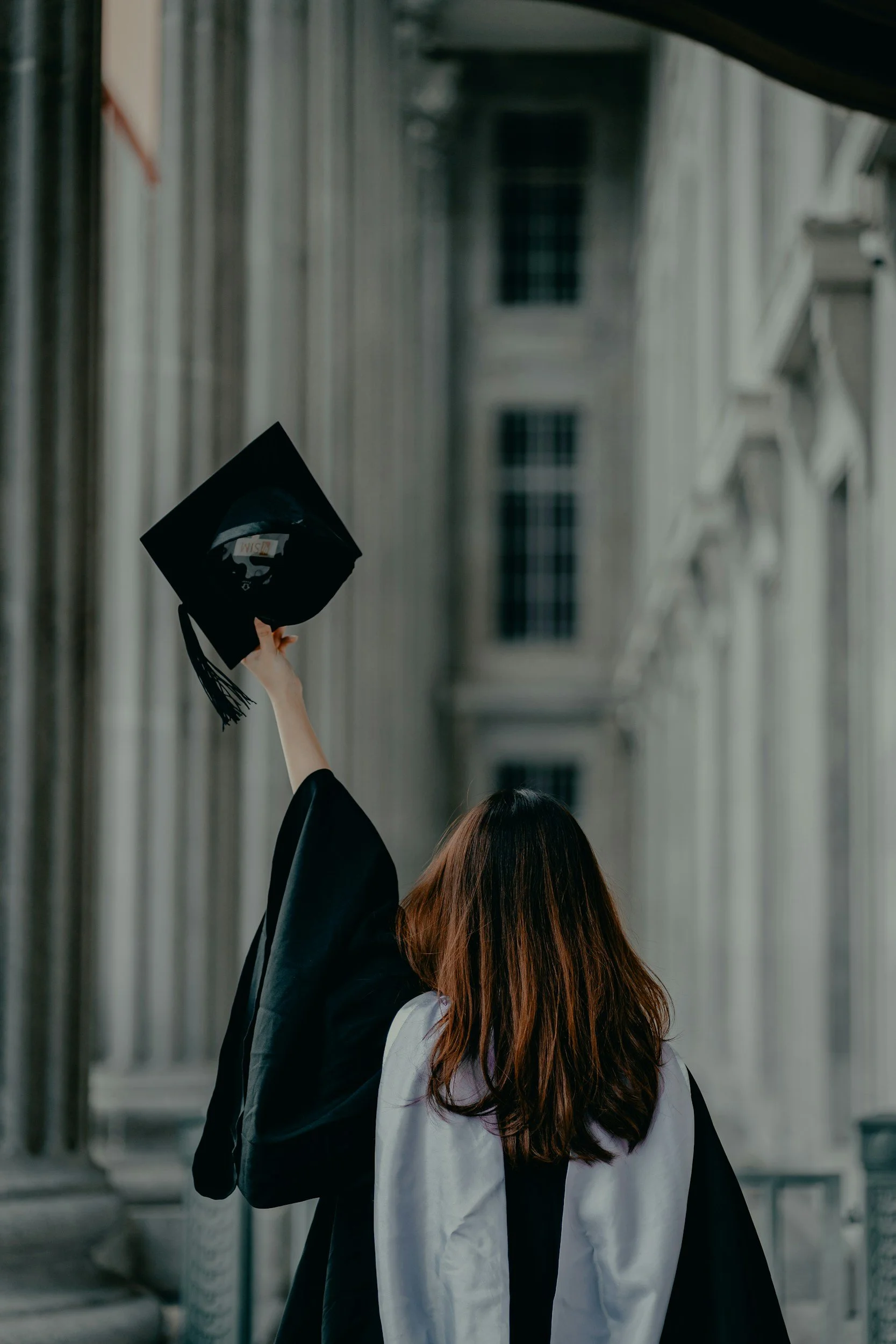 A woman in a graduation gown with a black and white stole holds her graduation cap high in the air outside a historic building with tall columns.