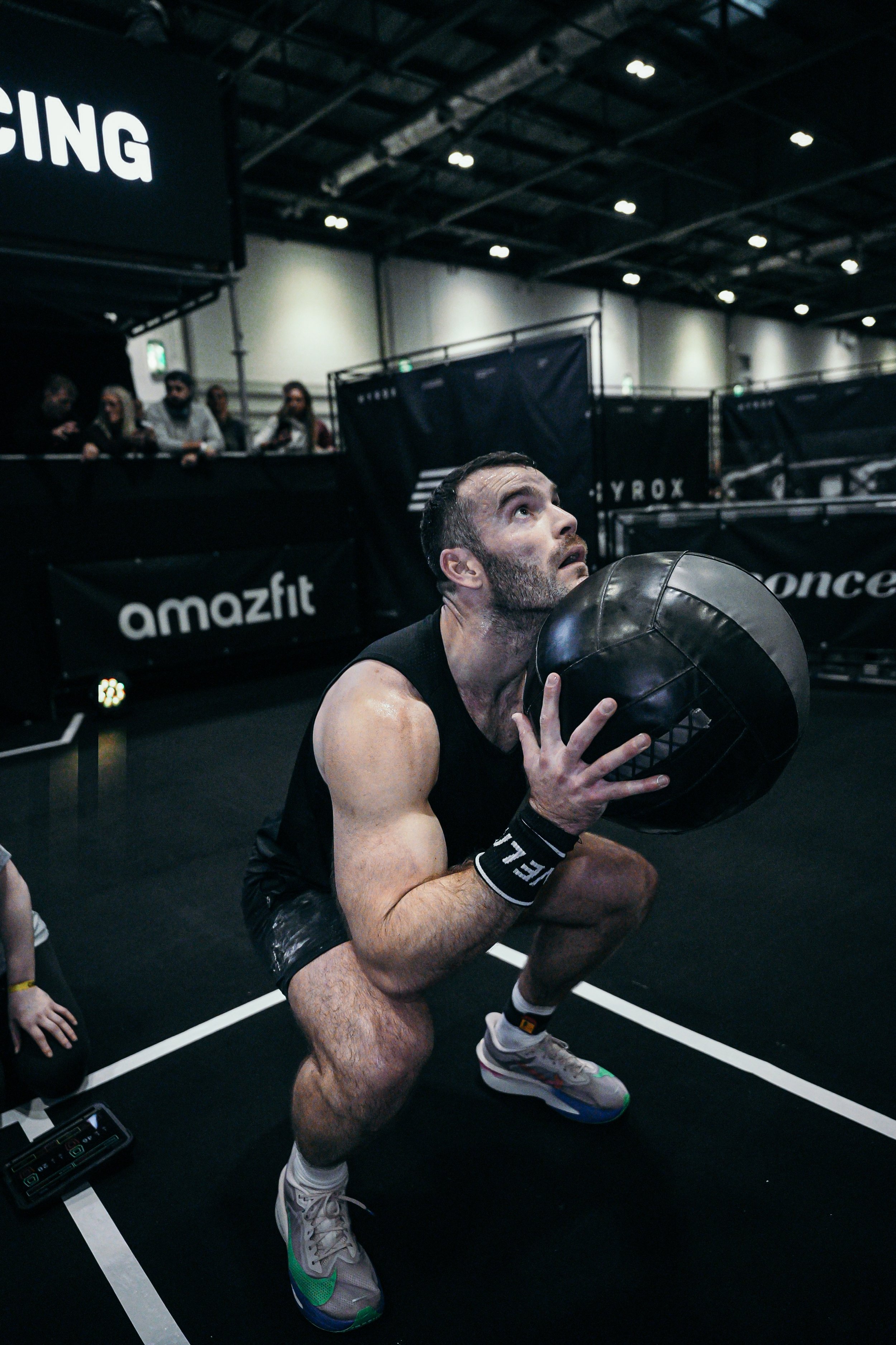 Dr Oliver  with short dark hair and a beard is squatting and holding a medicine ball with both hands during a workout or training session, in an indoor gym or fitness center with black flooring, black barriers, and spectators in the background.
